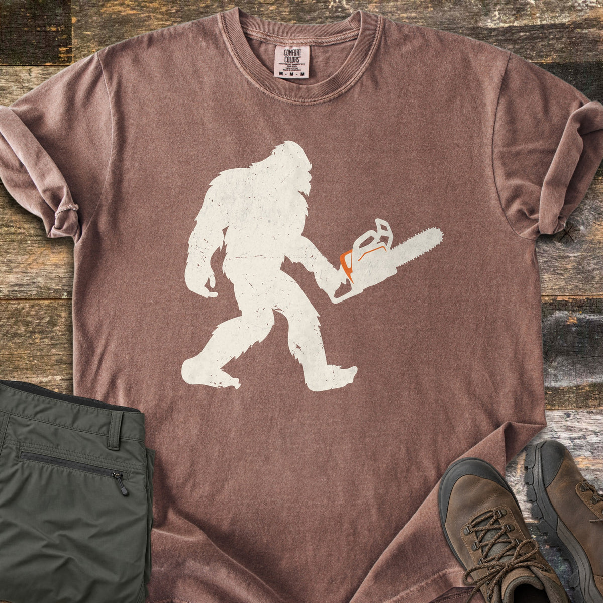 Lumber Squatch Comfort Colors T-shirt in Espresso, laid flat. A distressed cream silhouette of Bigfoot walks while carrying a chainsaw with orange accents. The sleeves are rolled, with folded olive pants and brown hiking boots on rustic wood planks.