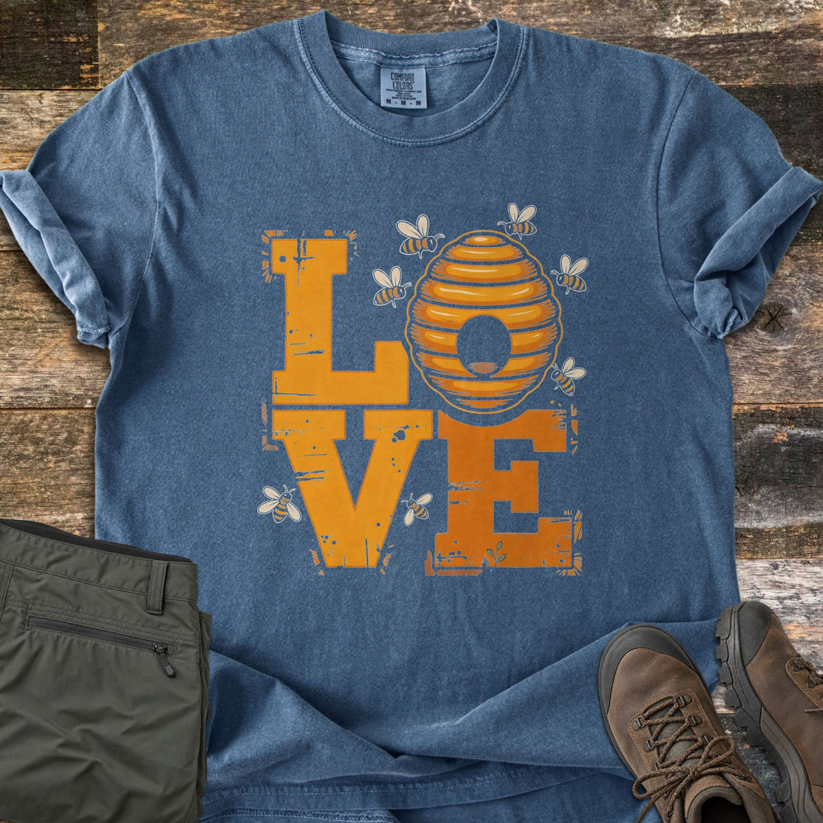 Beehive Love Comfort Colors T-shirt in Denim, laid flat. Distressed orange block letters spell LOVE, with a striped beehive as the O and small bees flying around it. Olive pants and brown hiking boots sit beside it on weathered wood planks.