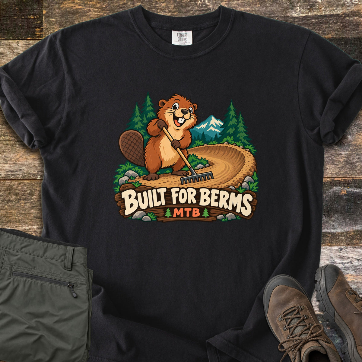 Built For Berms Comfort Colors T-shirt in Black, laid flat. A cartoon beaver holds a rake on a curved dirt berm, with pine trees and a snowy mountain behind, above a wooden sign reading Built For Berms MTB. Olive pants and brown hiking boots lie beside it on weathered wood.