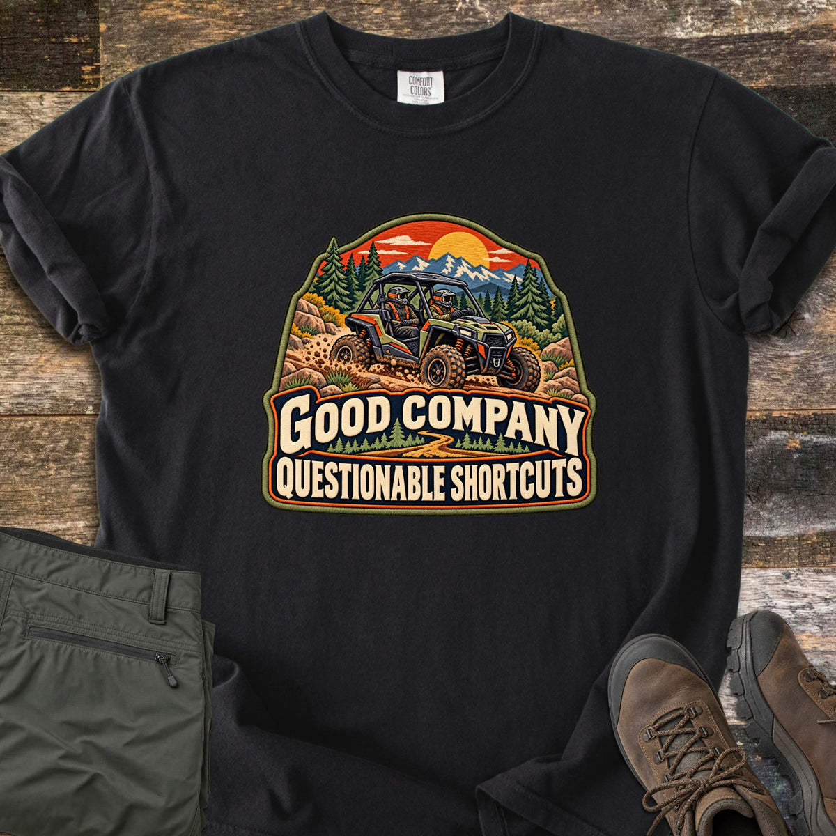 Good Company Questionable Shortcuts Comfort Colors T-shirt in Black, laid flat. An embroidered-style patch shows two riders in a side-by-side off-road vehicle kicking up dirt below mountains, pines and an orange sunset, above the words Good Company Questionable Shortcuts. Olive pants and brown boots sit on a wood floor.