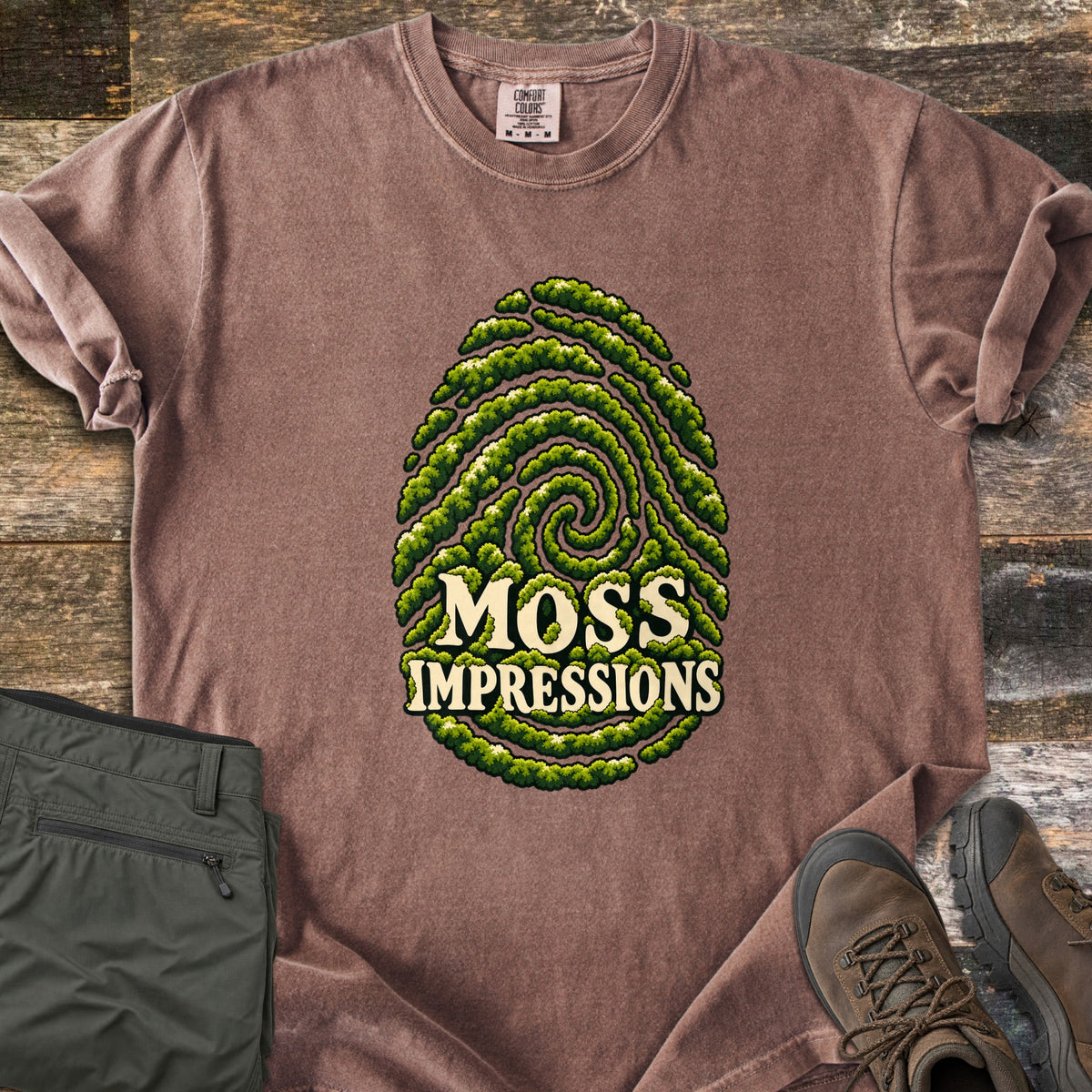 Moss Impressions Comfort Colors T-shirt in Espresso, laid flat. A fingerprint made of green moss ridges with the words Moss Impressions in cream letters. The shirt lies on weathered wood with folded olive hiking pants and brown hiking boots.
