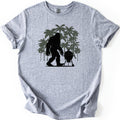 Squatch BBQ Smoker Comfort Colors T-shirt in Sport Grey, laid flat. A black silhouette of Bigfoot walks beside a kettle-style grill on wheeled legs, set against a cluster of olive green palm trees. No words are printed. The shirt lies on a white surface with sleeves cuffed.