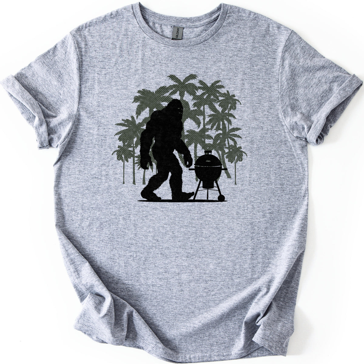 Squatch BBQ Smoker Comfort Colors T-shirt in Sport Grey, laid flat. A black silhouette of Bigfoot walks beside a kettle-style grill on wheeled legs, set against a cluster of olive green palm trees. No words are printed. The shirt lies on a white surface with sleeves cuffed.