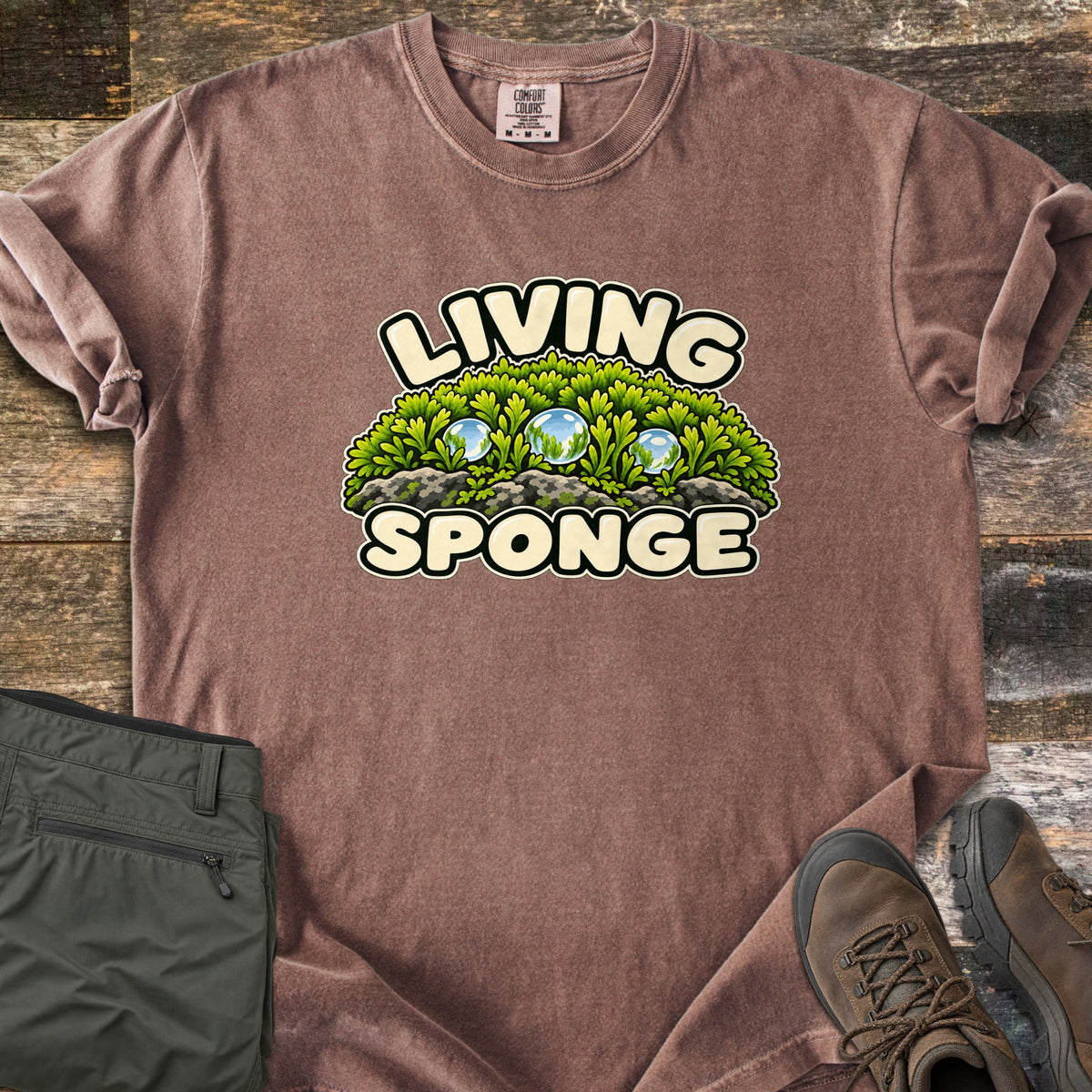 Living Sponge Comfort Colors T-shirt in Espresso, laid flat. A cartoon mound of bright green moss on gray rock, holding three clear water droplets, between the words Living Sponge. Olive hiking pants and brown leather hiking boots lie beside it on weathered wood.