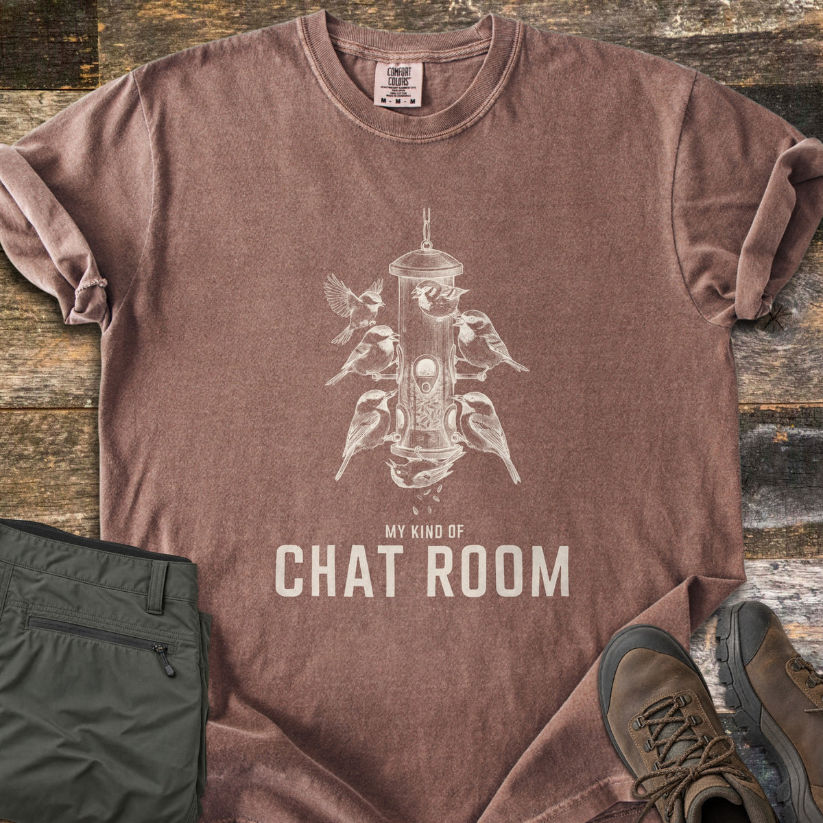 My Kind Of Chat Room Comfort Colors T-shirt in Espresso, laid flat. A cream sketch of chickadees perched around a hanging tube bird feeder, one upside down below it, above the words My Kind Of Chat Room. Olive hiking pants and brown boots lie beside it on weathered wood.