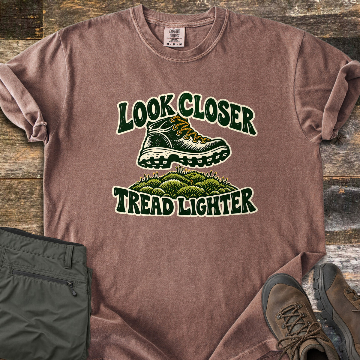Look Closer Tread Lighter Comfort Colors T-shirt in Espresso, laid flat. A green hiking boot with yellow laces steps above a mound of moss between the words Look Closer and Tread Lighter. Sleeves rolled, on weathered wood with folded olive hiking pants and brown boots.