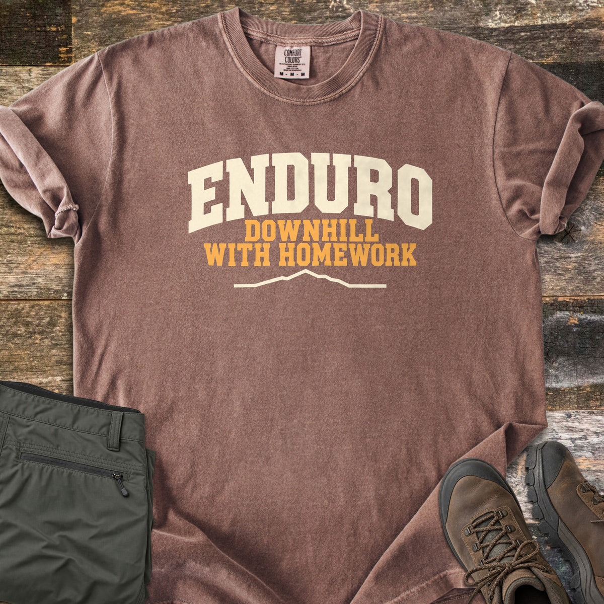 Enduro Downhill With Homework Comfort Colors T-shirt in Espresso, laid flat. The word Enduro in large cream arched letters, with Downhill With Homework in orange below and a thin cream mountain ridge line. Sleeves are rolled on a rustic wood surface beside green pants and brown hiking boots.