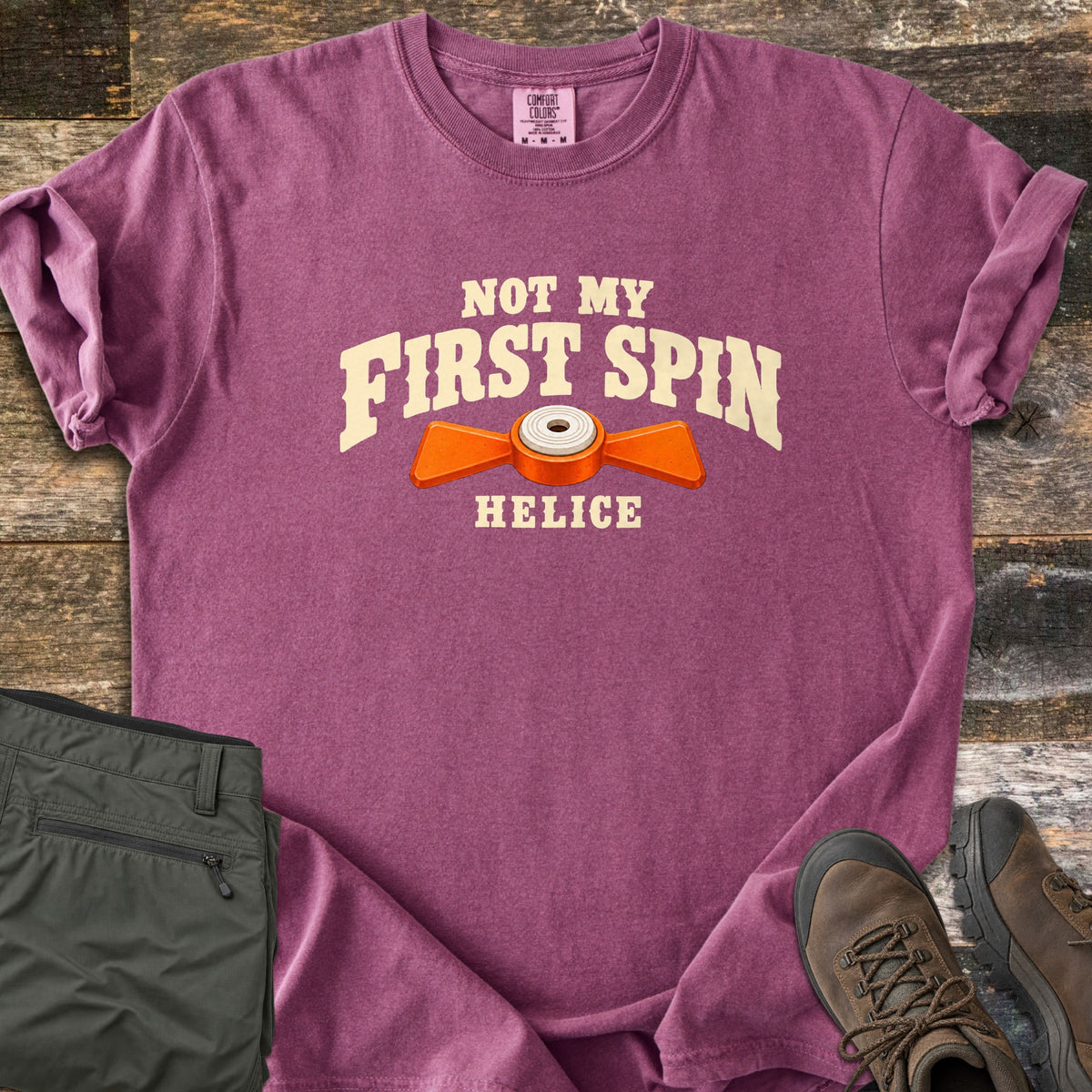 Not My First Spin Comfort Colors T-shirt in Berry, laid flat. Cream arched text reads Not My First Spin above an orange two-winged helice target with a white center, with Helice below. Folded olive pants and brown hiking boots sit beside it on weathered wood planks.