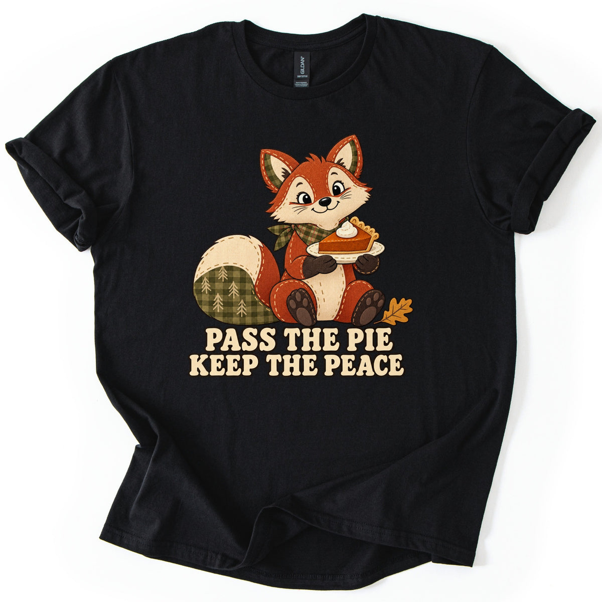 Pass the Pie Keep the Peace Comfort Colors T-shirt in Black, laid flat. A smiling patchwork fox in a green plaid bandana holds a slice of pumpkin pie with whipped cream above the words Pass the Pie Keep the Peace in cream letters. The shirt lies on a white background.
