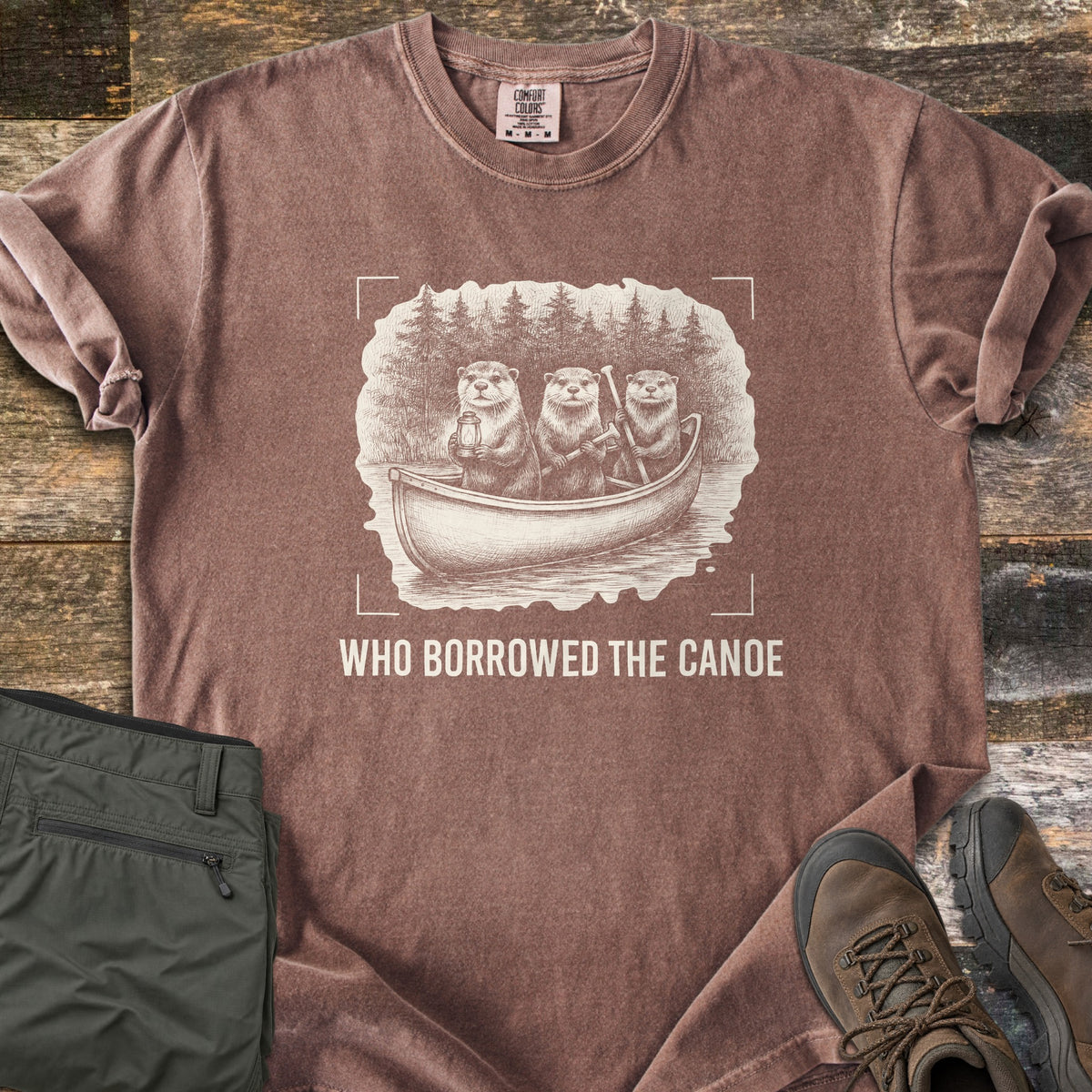 Who Borrowed The Canoe Comfort Colors T-shirt in Espresso, laid flat. A cream sketch of three otters in a canoe before pine trees, one holding a lantern, framed by camera corner marks above the words Who Borrowed The Canoe. Olive hiking pants and brown boots sit on wood.