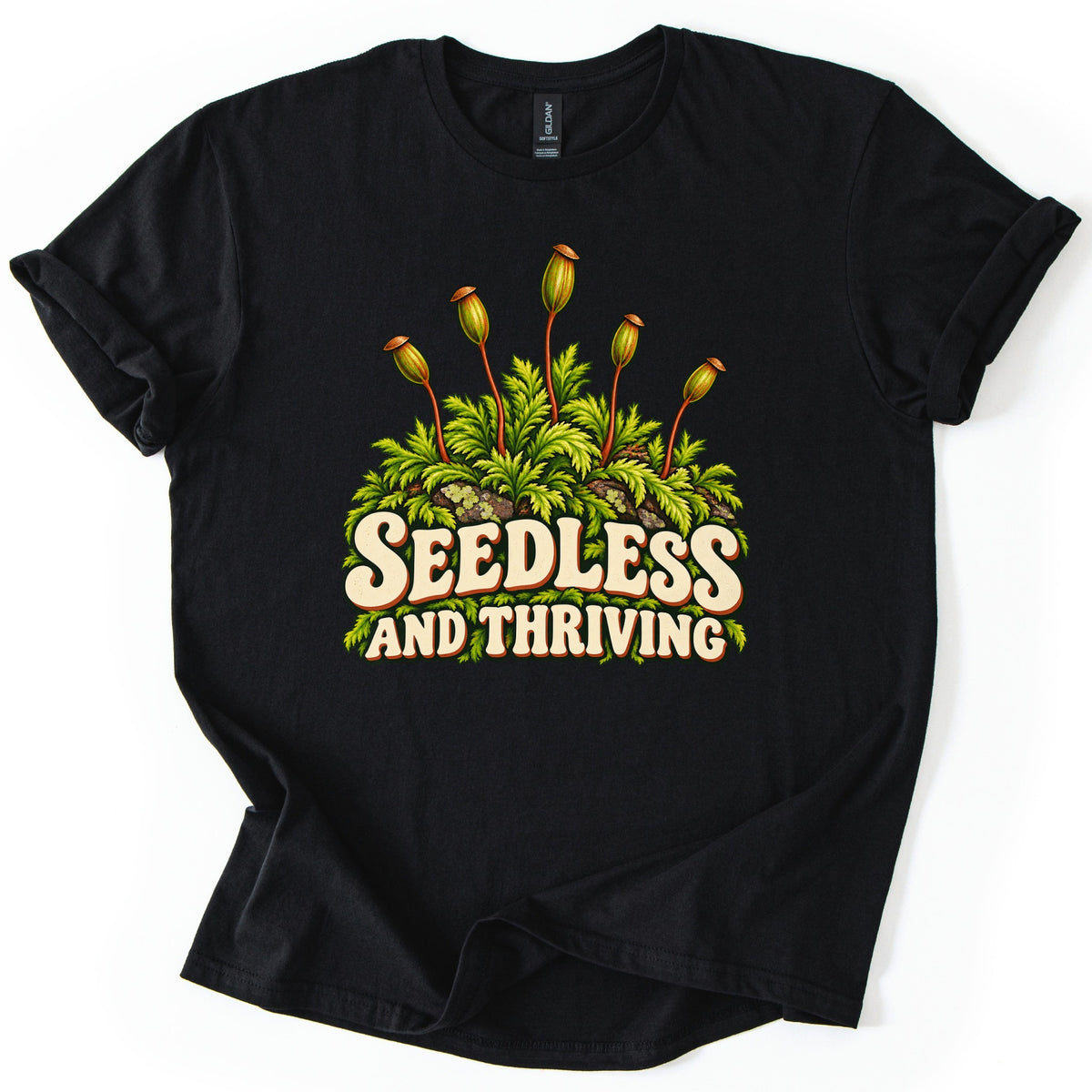 Seedless and Thriving Comfort Colors T-shirt in Black, laid flat. Green moss with tall reddish stalks topped by spore capsules grows over lichen-dotted rocks above the words SEEDLESS AND THRIVING in cream retro letters. The shirt lies on white with sleeves rolled.