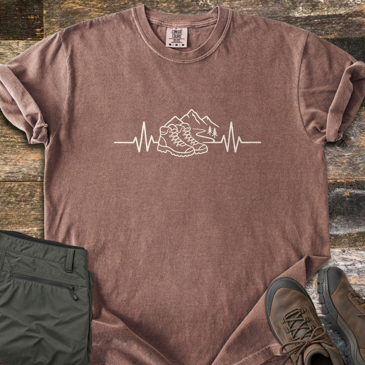 Hiking Heartbeat Comfort Colors T-shirt in Espresso, laid flat. A cream line drawing of hiking boots, mountains, a winding trail and two pine trees sits in a heartbeat line, with no words. Cuffed sleeves on weathered wood beside folded olive pants and brown boots.