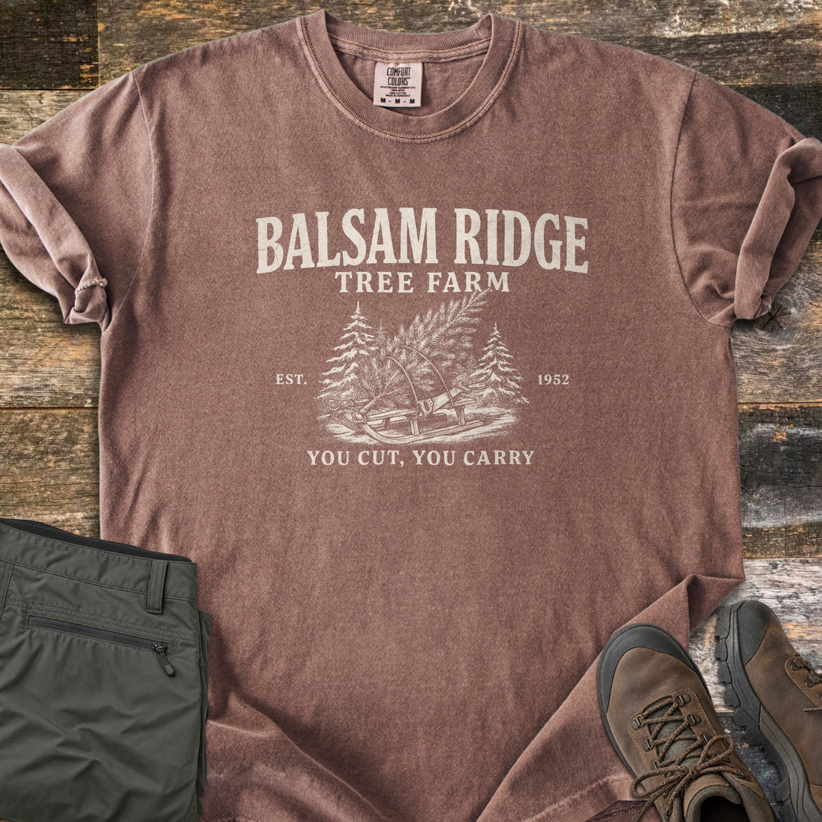 Balsam Ridge Tree Farm Comfort Colors T-shirt in Espresso, laid flat. Cream print reads Balsam Ridge Tree Farm above a cut evergreen tied to a wooden sled with a bow saw between two pines, Est. 1952, and You Cut, You Carry. Olive pants and brown hiking boots sit on weathered wood.