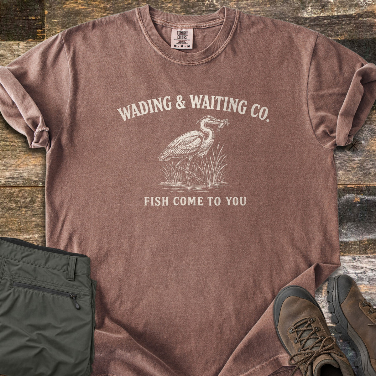 Wading & Waiting Co. Comfort Colors T-shirt in Espresso, laid flat. A heron stands in marsh reeds holding a fish in its beak, under the arched words Wading & Waiting Co. and above Fish Come To You. Olive cargo pants and brown hiking boots lie beside it on weathered wood.
