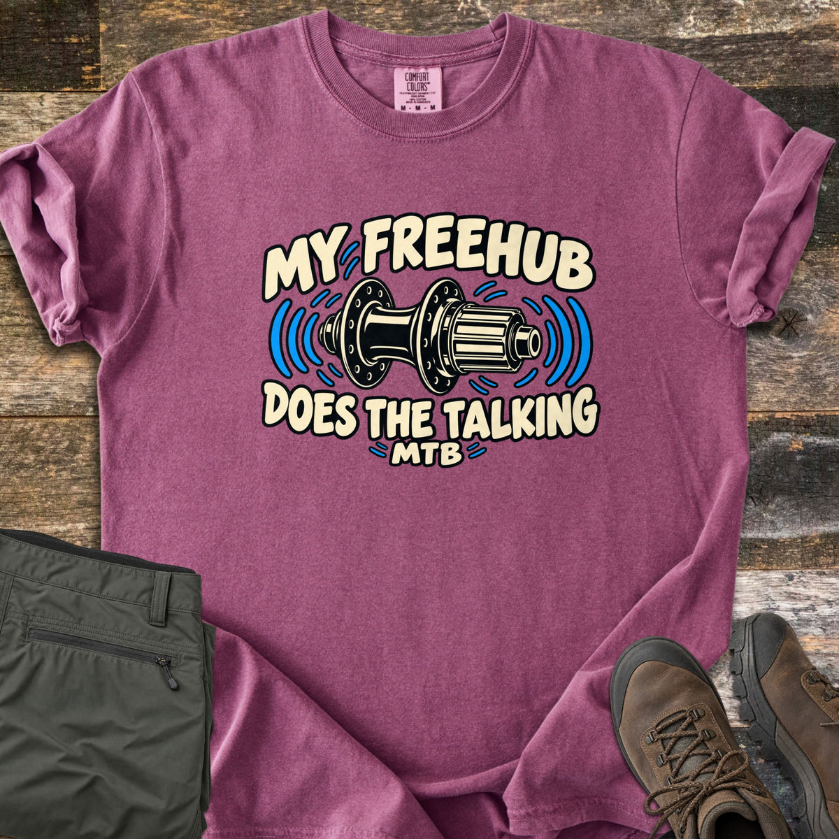 Freehub Does the Talking Comfort Colors T-shirt in Berry, laid flat. A black and cream bike rear hub with blue sound waves sits between the words My Freehub and Does the Talking, with MTB below. Olive hiking pants and brown boots lie beside it on weathered wood.