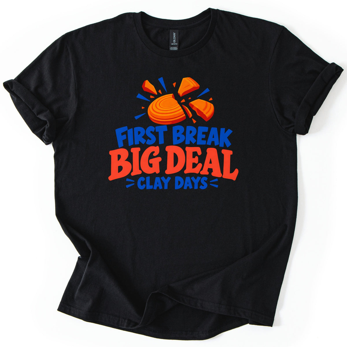 First Break Big Deal Comfort Colors T-shirt in Black, laid flat. An orange clay target breaking apart with blue burst lines sits above the words First Break Big Deal Clay Days, in blue and red-orange letters. The sleeves are rolled on a plain white background.