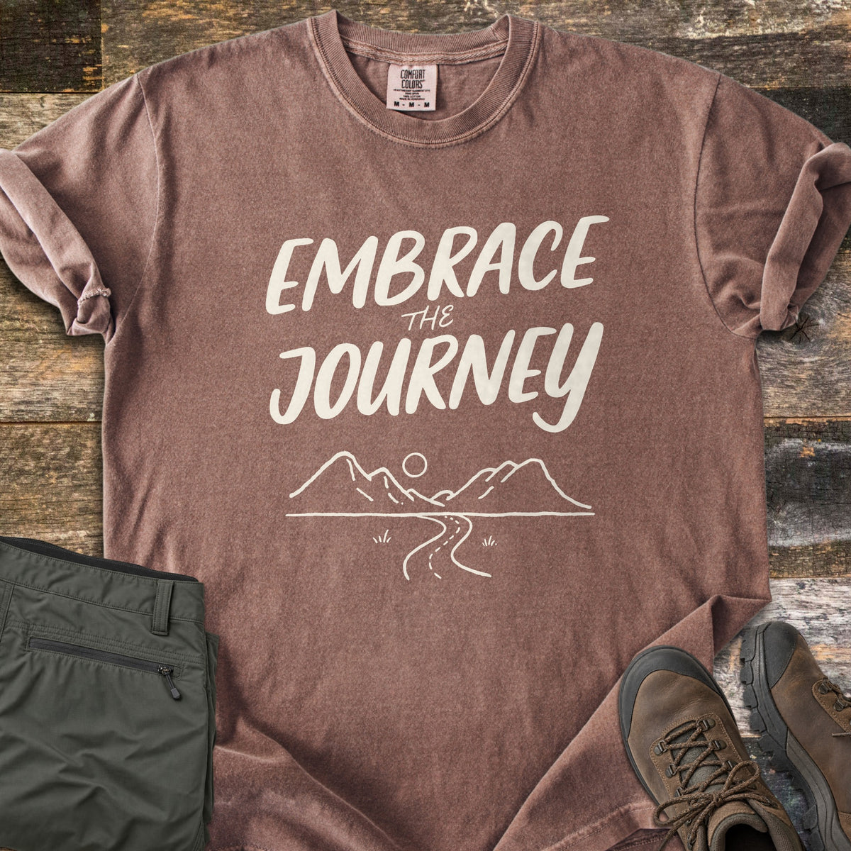 Embrace the Journey Comfort Colors T-shirt in Espresso, laid flat. Cream brush lettering reads Embrace the Journey above a line drawing of mountains, a sun, and a winding road. Sleeves cuffed on weathered wood beside folded green pants and brown hiking boots.