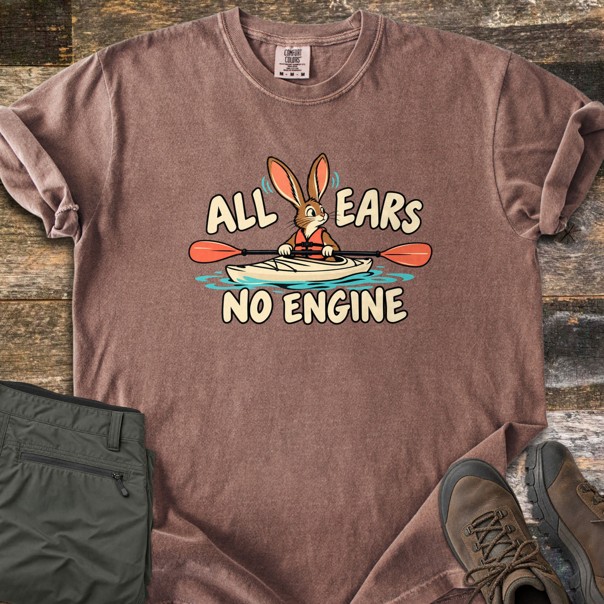 All Ears No Engine Comfort Colors T-shirt in Espresso, laid flat. A cartoon rabbit in an orange life vest paddles a cream kayak with an orange-bladed paddle on blue water, between the words All Ears No Engine. Olive cargo pants and brown hiking boots sit on a wood plank floor.