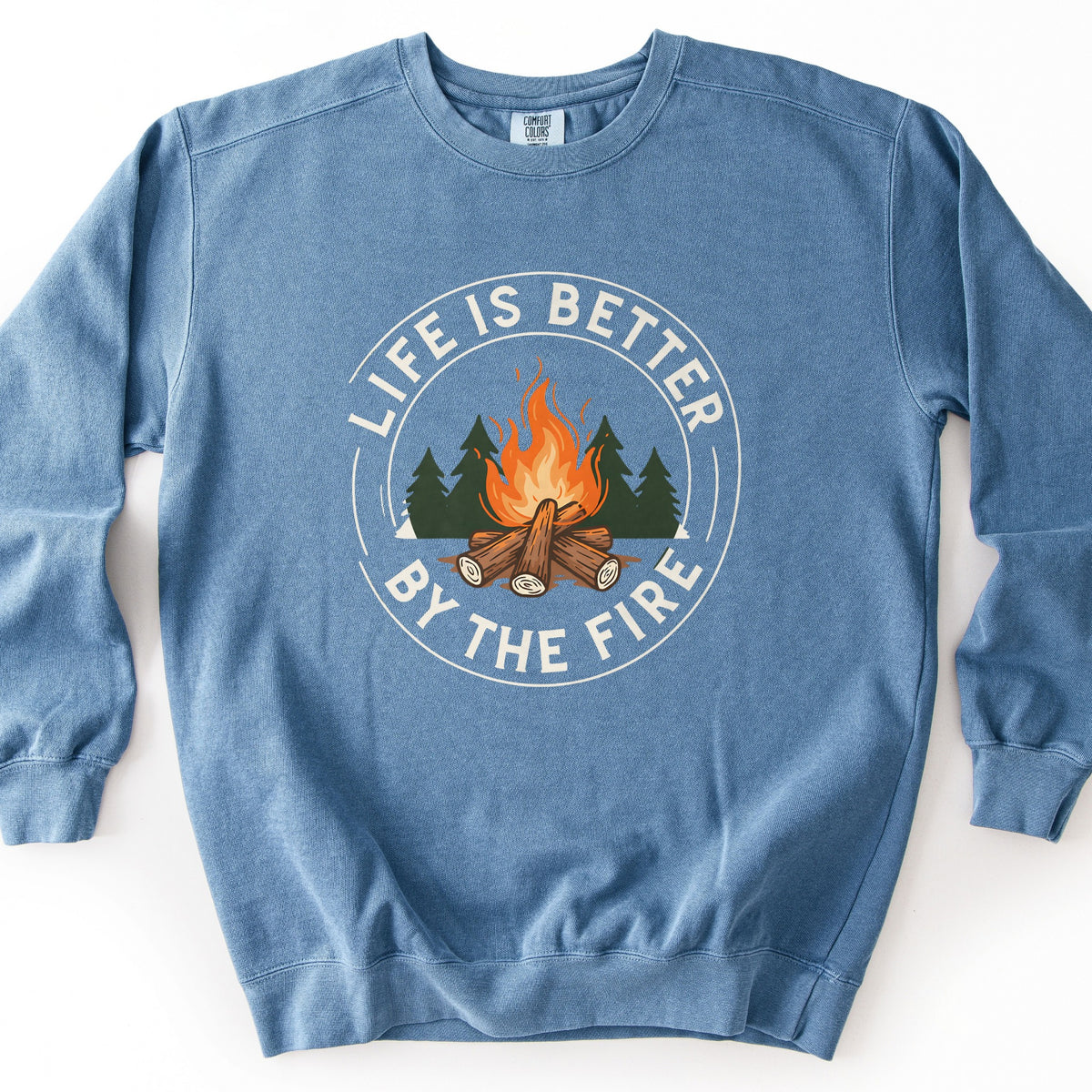 Life Is Better By The Fire Ring Comfort Colors crewneck sweatshirt in Blue Jean, laid flat. A circular badge shows a campfire of crossed logs with orange flames before dark green pine trees, ringed by the words Life Is Better By The Fire in cream lettering. It lies on a white surface.
