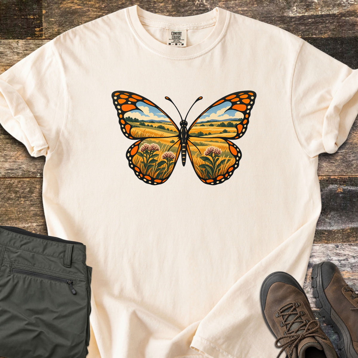 Monarch Prairie Comfort Colors T-shirt in Ivory, laid flat. A monarch butterfly whose wings frame a prairie scene of golden grass, pink milkweed blooms and a blue sky with clouds. Olive cargo pants and brown hiking boots sit beside it on weathered wood planks.