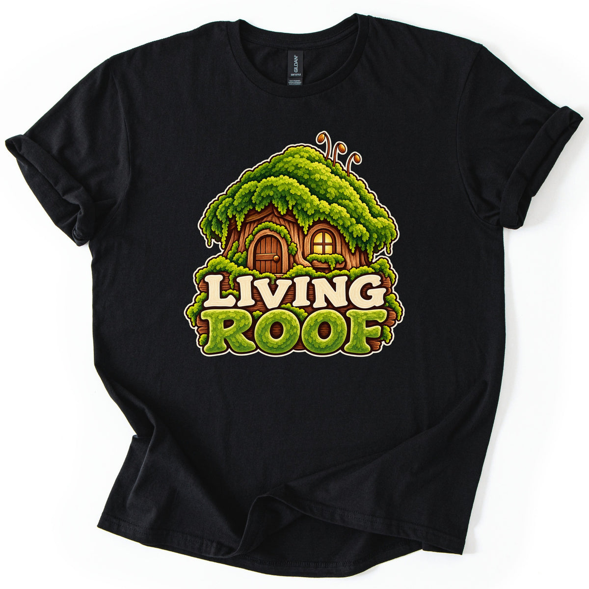 Living Roof Comfort Colors T-shirt in Black, laid flat. A cartoon tree-stump house topped with a heavy green moss roof and two curled sprouts, with an arched door and lit window, above the words Living Roof. The shirt lies on a white surface with cuffed sleeves.