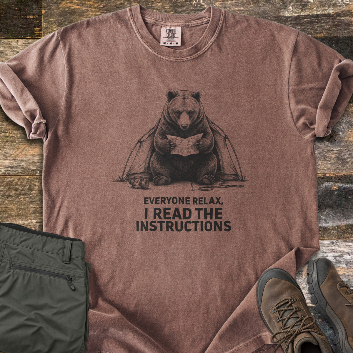 Everyone Relax I Read The Instructions Comfort Colors T-shirt in Espresso, laid flat. A black sketch of a bear reading instructions in front of a tent, with a tipped lantern and rope, above the words Everyone relax, I read the instructions. Olive cargo pants and brown hiking boots sit on dark wood.