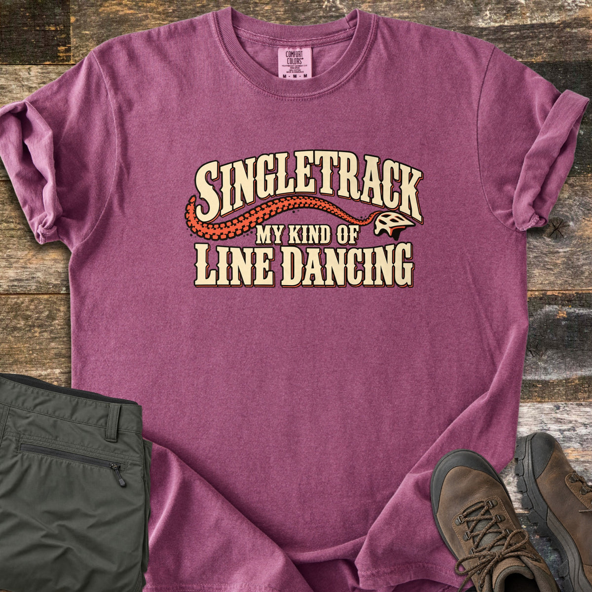 Singletrack My Kind of Line Dancing Comfort Colors T-shirt in Berry, laid flat. Cream western-style lettering reads Singletrack, My Kind of Line Dancing, with an orange tire track curving to a bike helmet. Shown on weathered wood with folded green hiking pants and brown hiking boots.