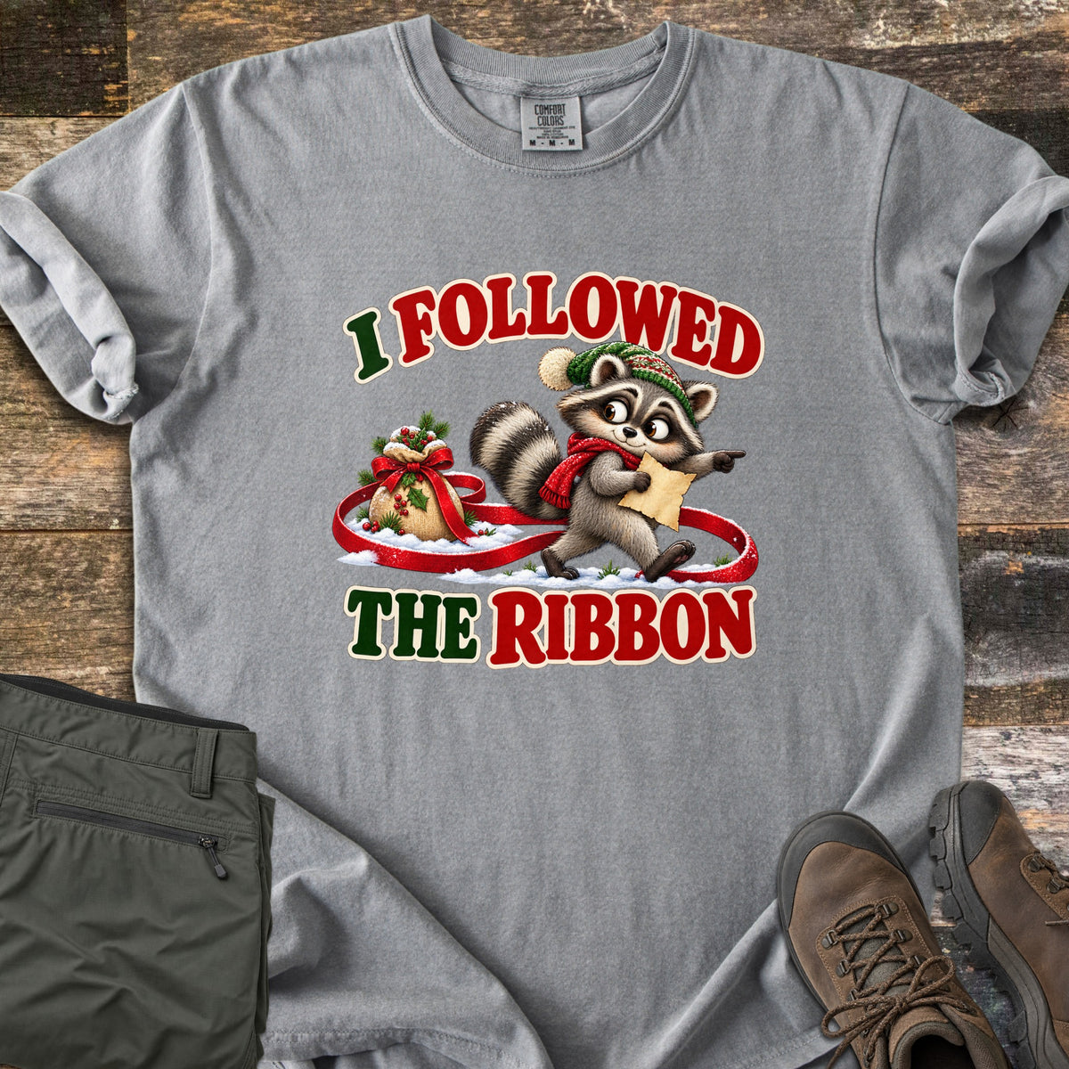 I Followed The Ribbon Comfort Colors T-shirt in Grey, laid flat. A cartoon raccoon in a green knit hat and red scarf holds a map and points ahead, walking along a red ribbon from a gift sack in snow, between the words I Followed The Ribbon. Olive pants and brown boots lie on a wood floor.