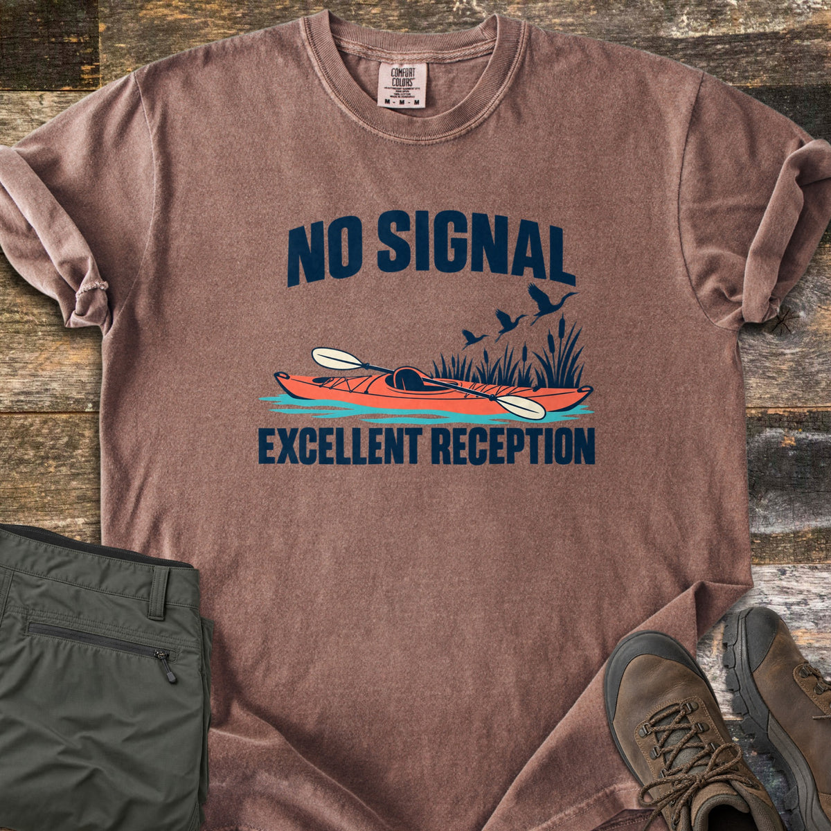 Excellent Reception Comfort Colors T-shirt in Espresso, laid flat. The words No Signal arch over an orange kayak with a paddle among reeds and flying birds, above the words Excellent Reception. Olive pants and brown hiking boots sit on a weathered wood surface.