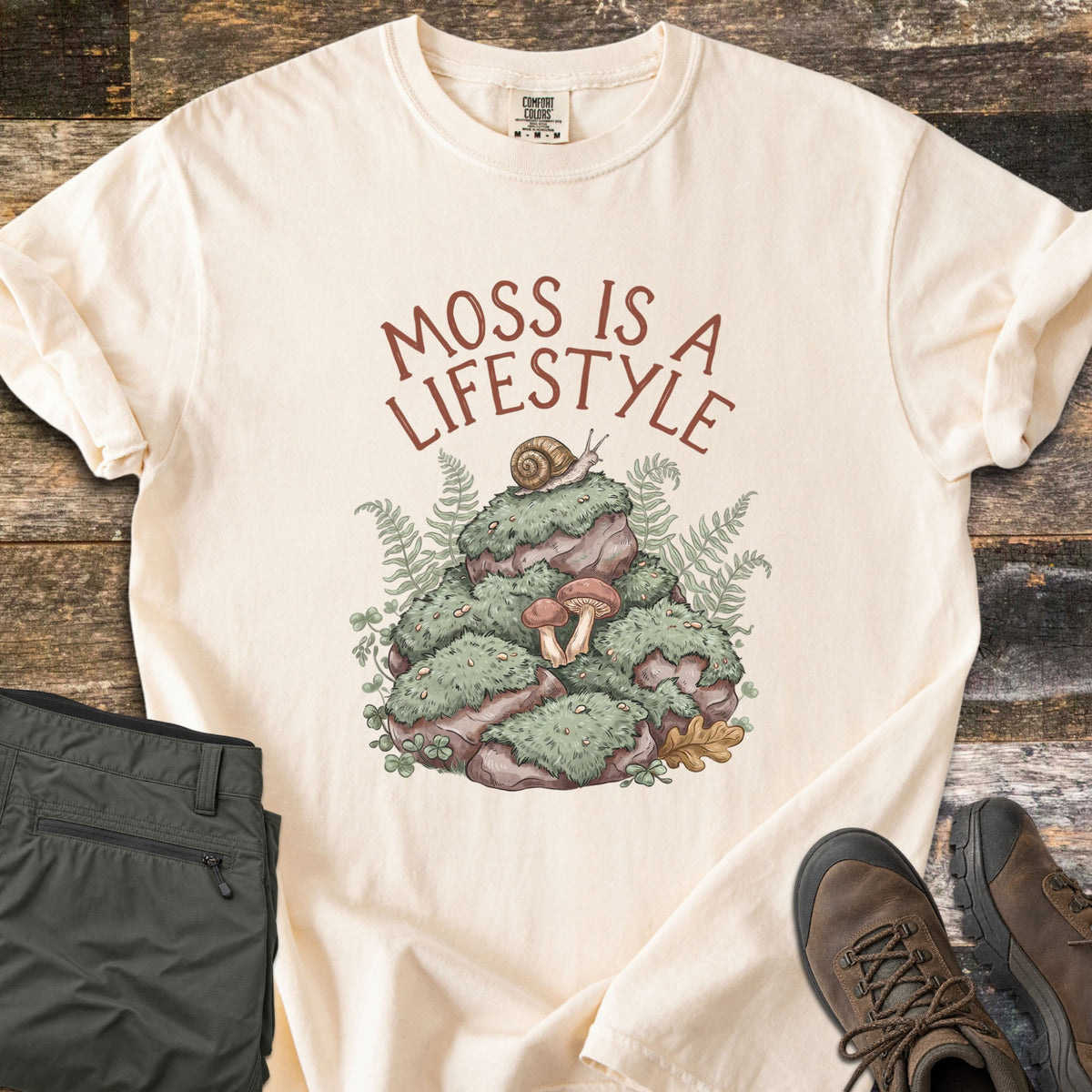 Moss Is A Lifestyle Comfort Colors T-shirt in Ivory, laid flat. Rust-red text reads Moss Is A Lifestyle above a mossy rock mound with a snail, mushrooms, ferns and clover. It lies on weathered wood beside folded olive hiking pants and brown hiking boots.