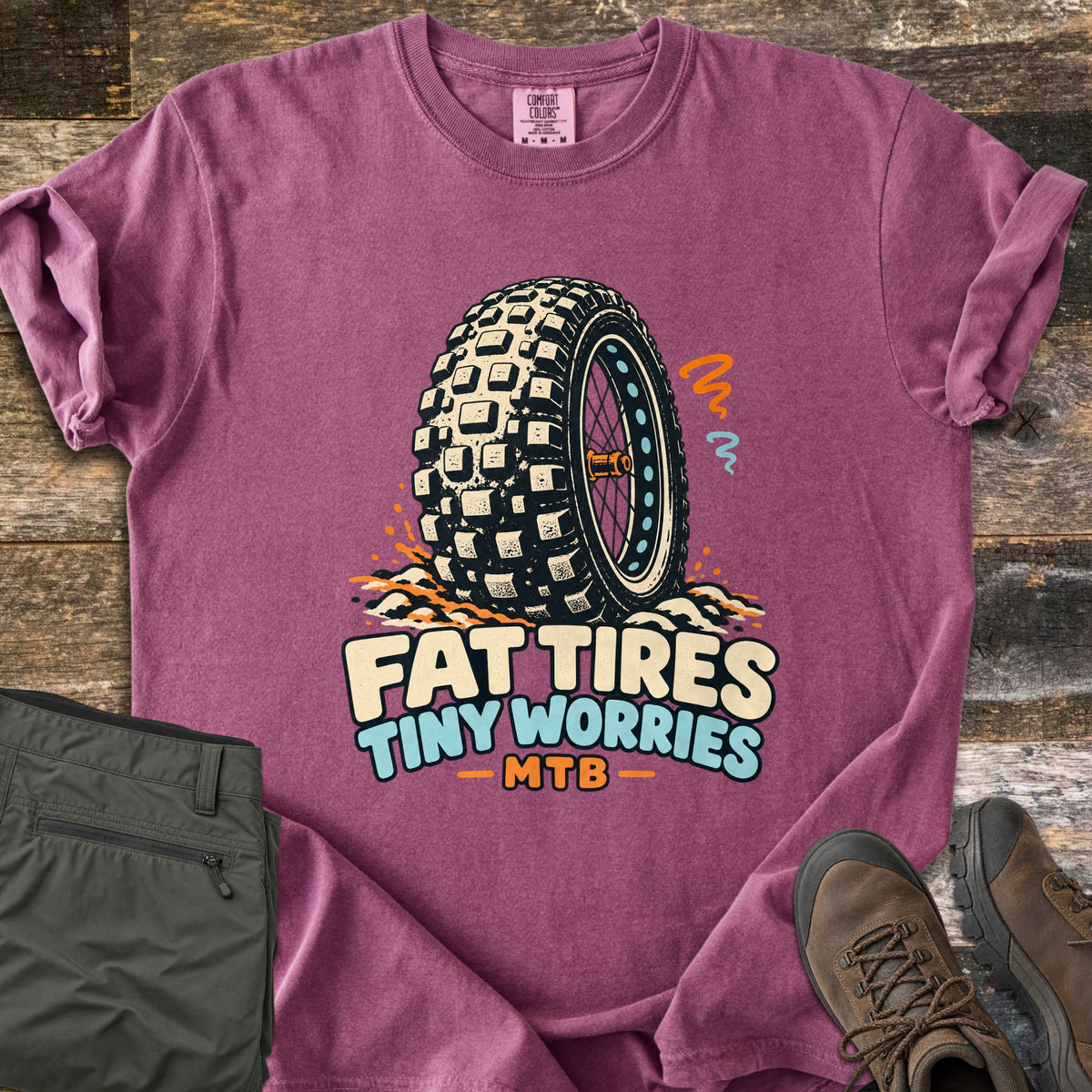 Fat Tires Tiny Worries Comfort Colors T-shirt in Berry, laid flat. A knobby fat bike tire kicking up dirt, with orange and blue squiggles, above the words Fat Tires Tiny Worries MTB. Sleeves rolled, on weathered wood with olive pants and brown hiking boots.