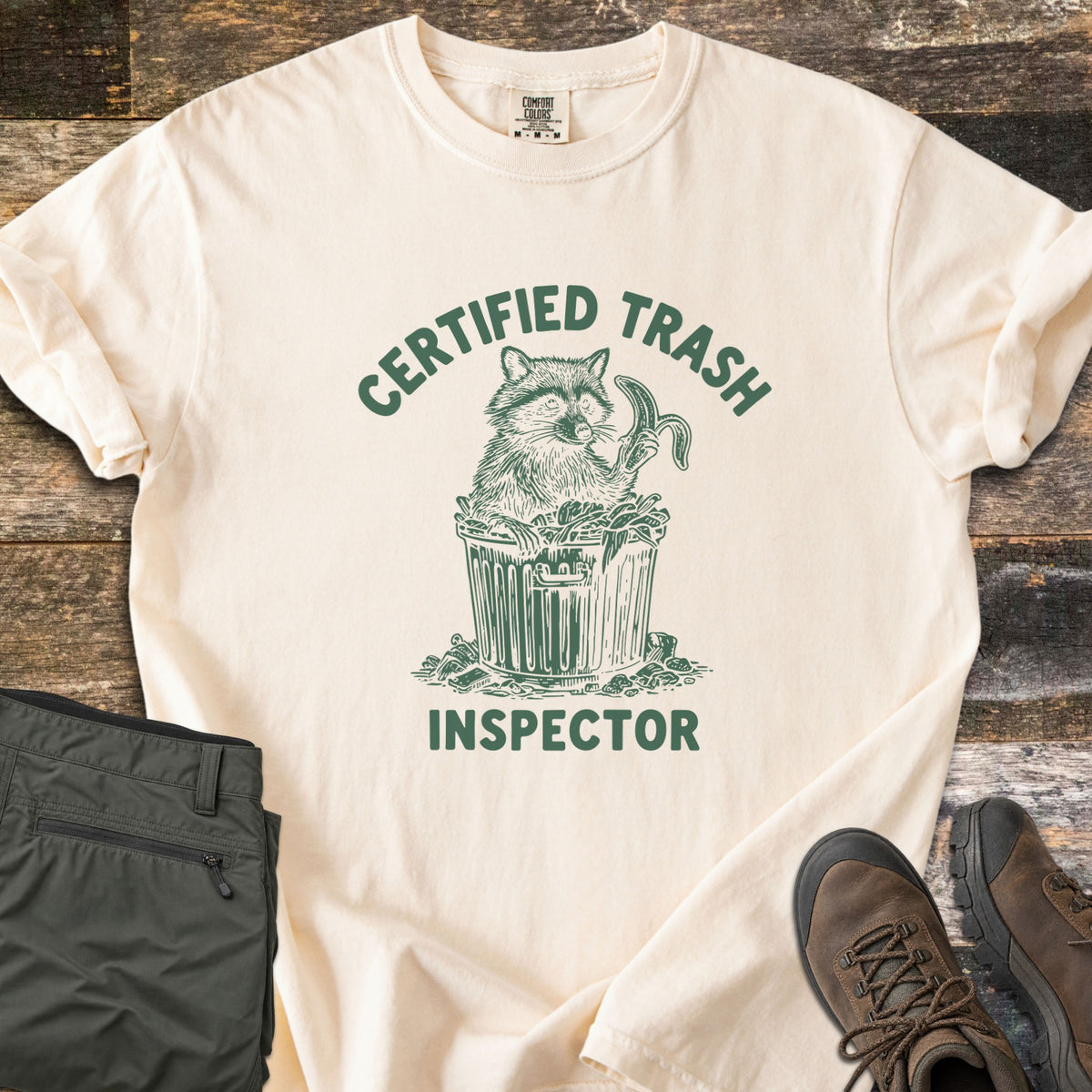Certified Trash Inspector Comfort Colors T-shirt in Ivory, laid flat. A green sketch of a raccoon in an overflowing trash can holding a banana peel, with the words Certified Trash arched above and Inspector below. Olive cargo pants and brown hiking boots lie on a wood floor.