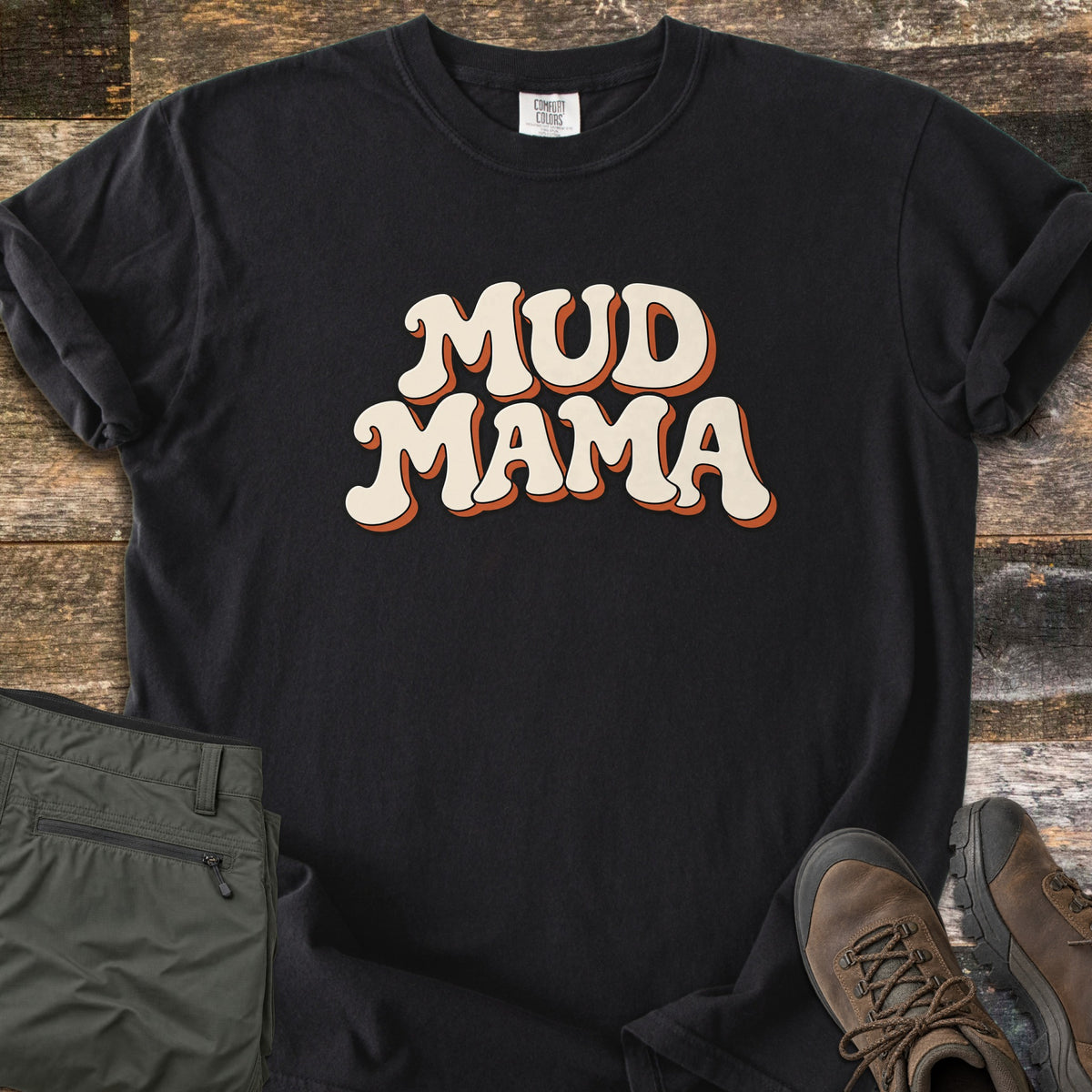 Mud Mama Comfort Colors T-shirt in Black, laid flat. The words MUD MAMA are printed in cream retro bubble letters with an orange drop shadow. The shirt lies on weathered wood planks beside folded olive hiking pants and brown hiking boots.
