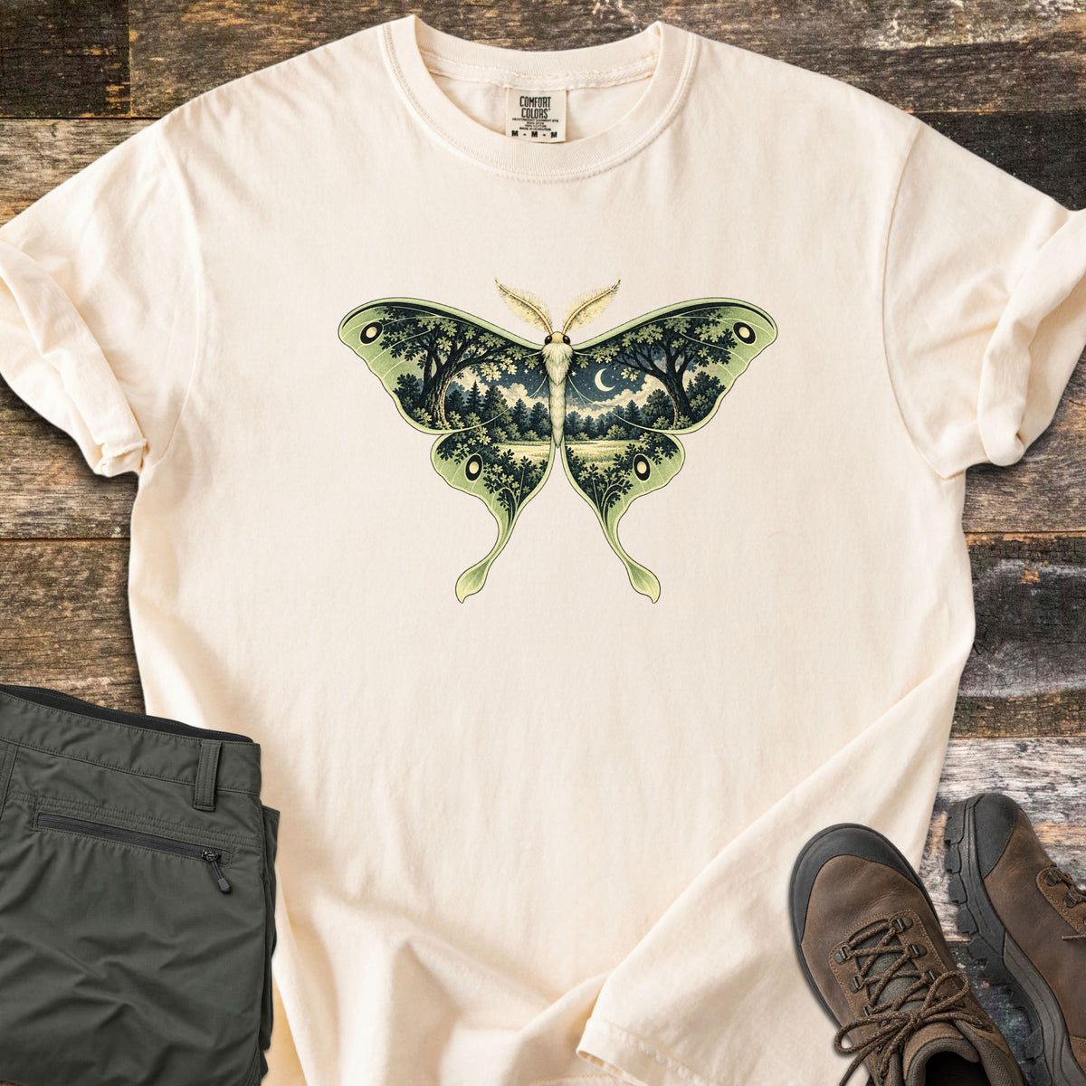 Luna Moth Night Comfort Colors T-shirt in Ivory, laid flat. A luna moth with pale green wings, each wing filled with a night forest scene of dark trees, a meadow and a crescent moon. Rolled sleeves; olive cargo pants and brown hiking boots lie beside it on weathered wood.