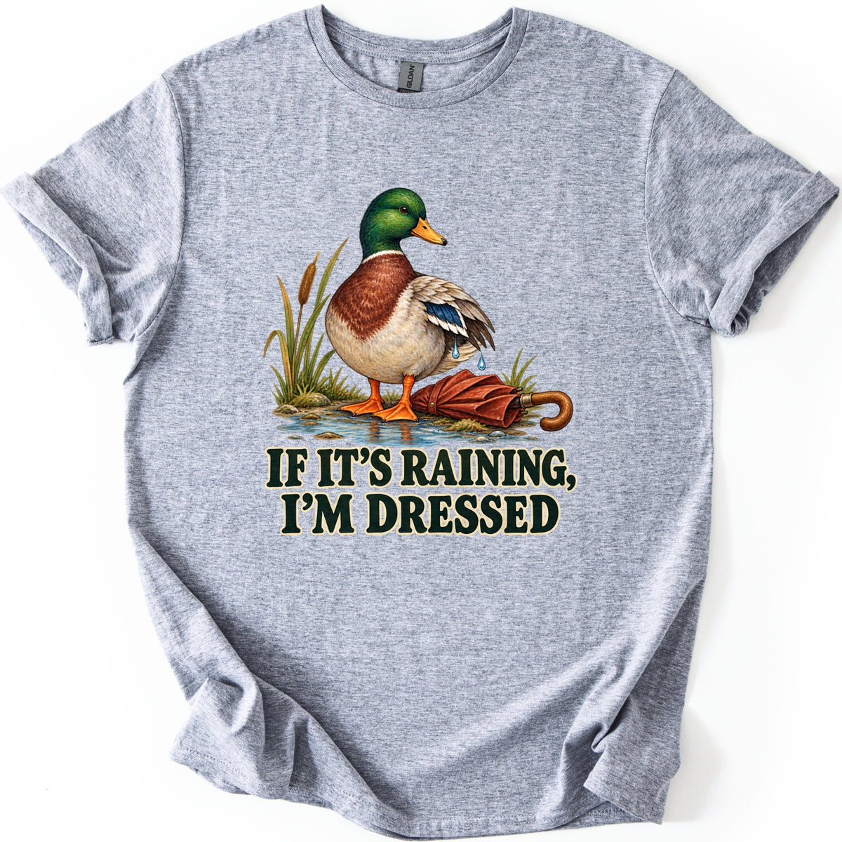 If It's Raining, I'm Dressed Comfort Colors T-shirt in Sport Grey, laid flat. A mallard duck with water dripping off its wing stands in a puddle by cattails beside a closed red umbrella, above the words IF IT'S RAINING, I'M DRESSED in dark green. Cuffed sleeves, white background.