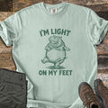 I'm Light On My Feet Comfort Colors T-shirt in Bay, laid flat. A green etched illustration of a round hippo dancing on its hind legs, with I'M LIGHT above and ON MY FEET below. Sleeves are rolled on a dark wood surface beside folded olive pants and brown hiking boots.