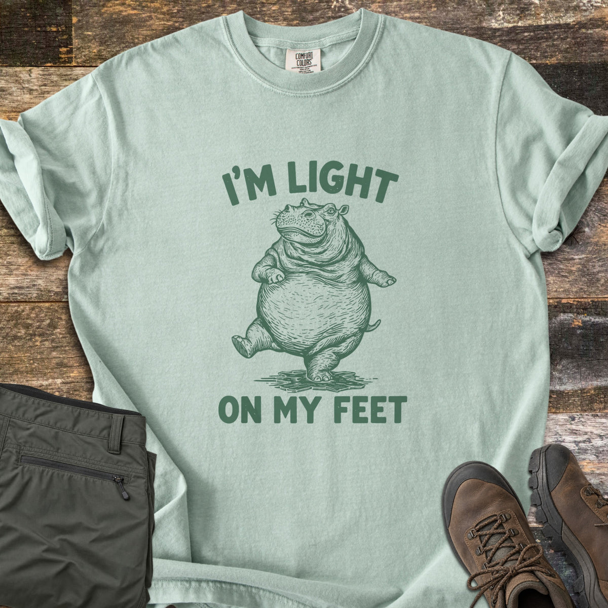 I'm Light On My Feet Comfort Colors T-shirt in Bay, laid flat. A green etched illustration of a round hippo dancing on its hind legs, with I'M LIGHT above and ON MY FEET below. Sleeves are rolled on a dark wood surface beside folded olive pants and brown hiking boots.