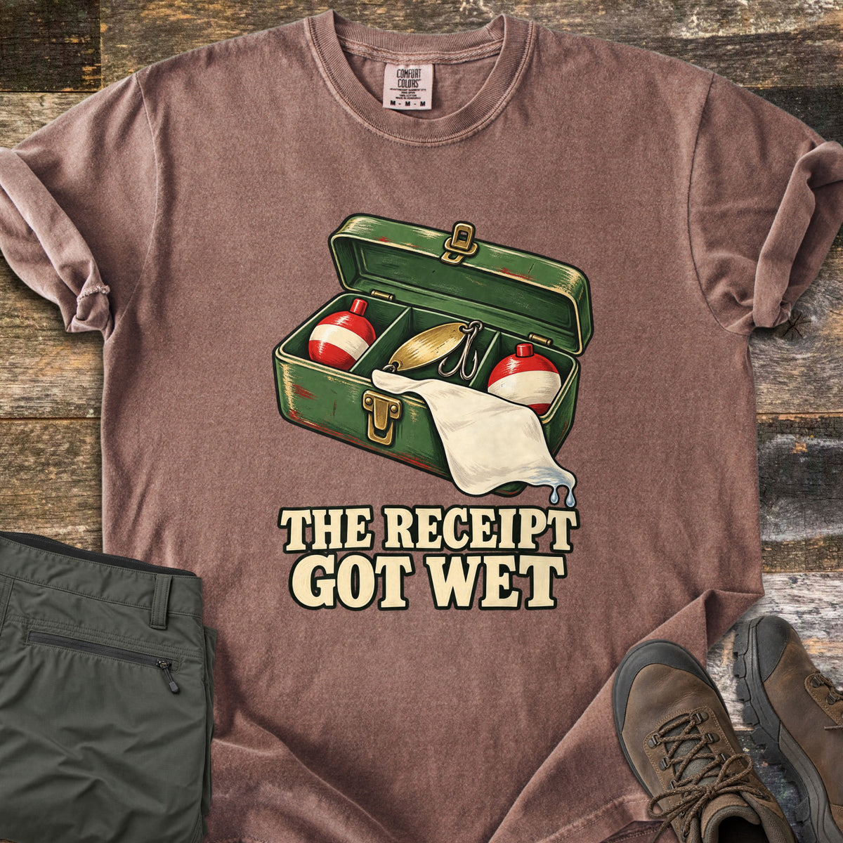 The Receipt Got Wet Comfort Colors T-shirt in Espresso, laid flat. An open green tackle box holds red and white bobbers and a gold spoon lure, with a dripping white paper hanging out, above the words The Receipt Got Wet. Olive pants and brown hiking boots sit on weathered wood.