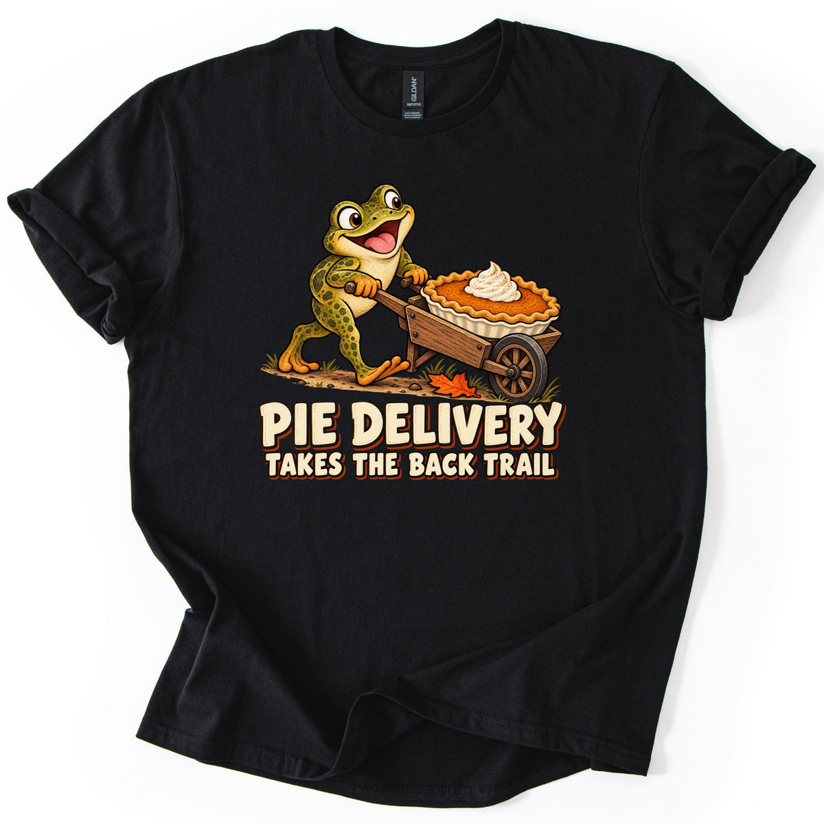 Pie Delivery Takes the Back Trail Comfort Colors T-shirt in Black, laid flat. A grinning cartoon frog pushes a wooden wheelbarrow carrying a whipped-cream-topped pumpkin pie past an autumn leaf, above the words Pie Delivery Takes the Back Trail. Rolled sleeves on a white background.