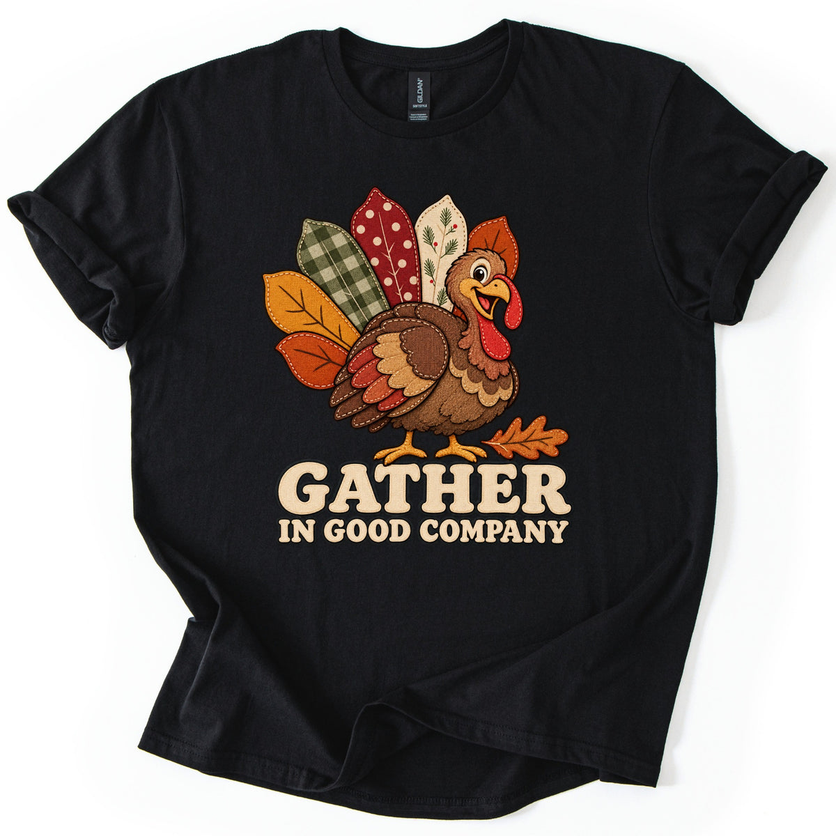 Gather in Good Company Comfort Colors T-shirt in Black, laid flat. A cheerful cartoon turkey with quilted tail feathers in green gingham, red polka dot and fall leaf patterns stands above the words GATHER IN GOOD COMPANY in cream letters. Sleeves rolled, on a white surface.