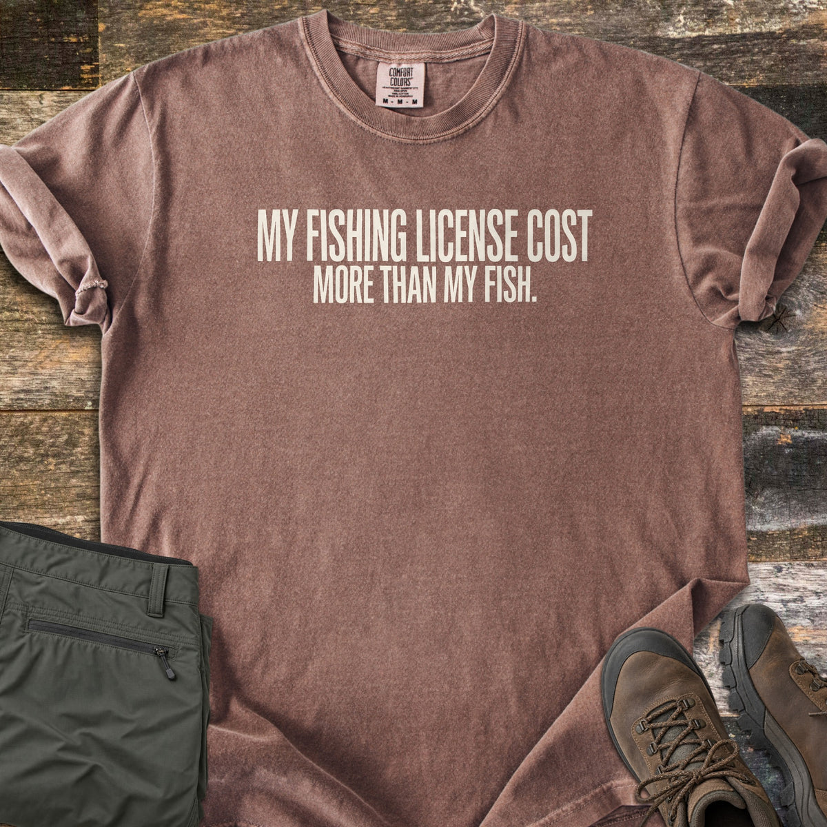 License Cost More Than My Fish Comfort Colors T-shirt in Espresso, laid flat. Cream block letters read My fishing license cost more than my fish. The shirt has rolled sleeves and lies on weathered wood planks beside folded olive hiking pants and brown hiking boots.