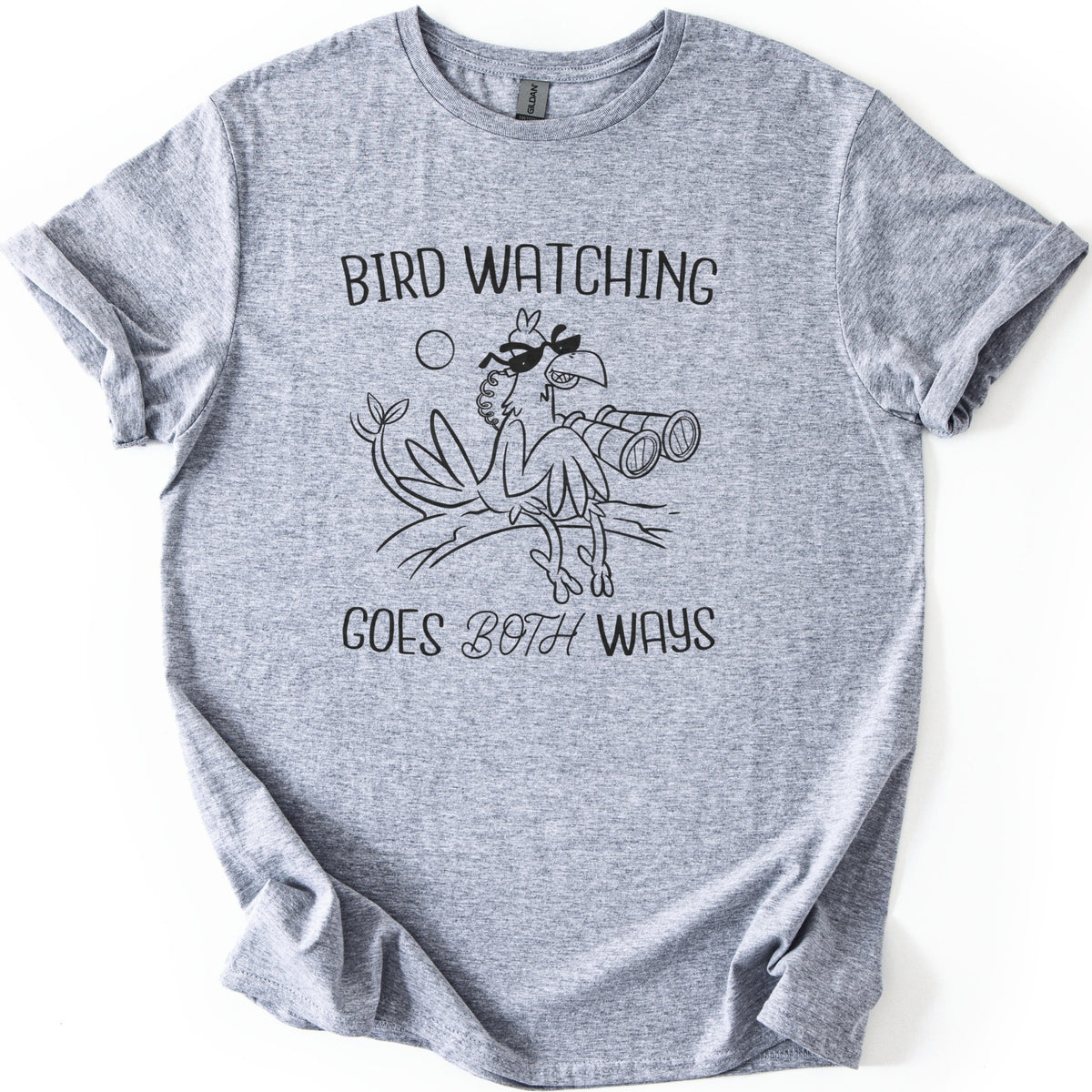 Birds Watch Back Birder Comfort Colors T-shirt in Sport Grey, laid flat. Black line art of a cartoon bird in sunglasses perched on a branch beside a pair of binoculars, with a small sun, between the words Bird Watching Goes Both Ways. Sleeves rolled, on a white background.