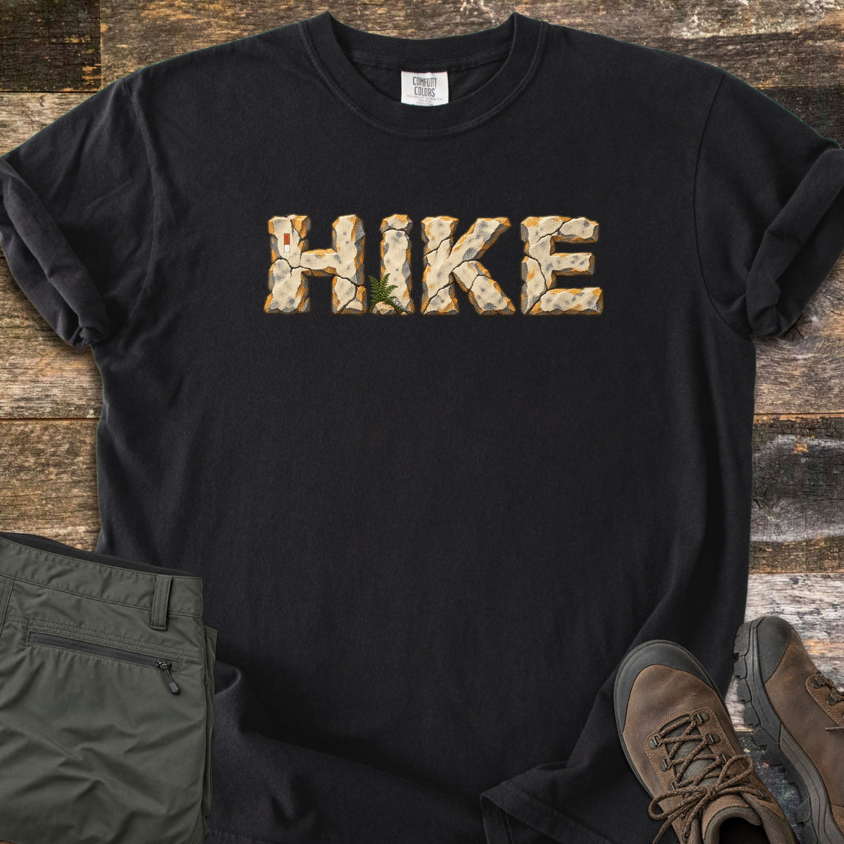 Hike Comfort Colors T-shirt in Black, laid flat. The word HIKE is printed across the chest in cracked stone letters with orange edges, a small fern at the base of the I. Olive hiking pants and brown hiking boots lie beside it on weathered wood planks.