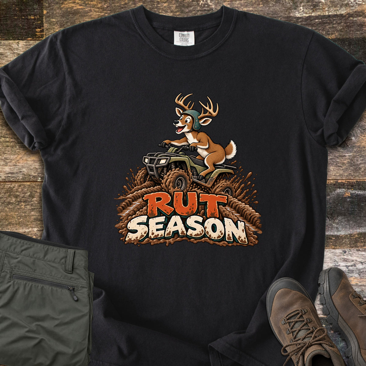 Rut Season Comfort Colors T-shirt in Black, laid flat. A cartoon buck in a green helmet rides a four-wheeler through splashing mud above the words Rut Season. Sleeves are rolled on a wood plank surface beside folded olive pants and brown hiking boots.