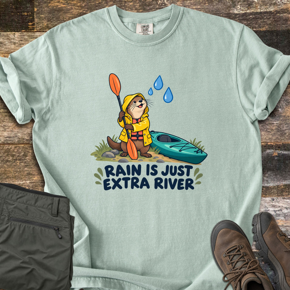 Extra River Comfort Colors T-shirt in Bay, laid flat. A cartoon otter in a yellow raincoat and life vest holds an orange paddle beside a teal kayak as raindrops fall, above the words Rain Is Just Extra River. Olive pants and brown hiking boots lie on a wood floor.