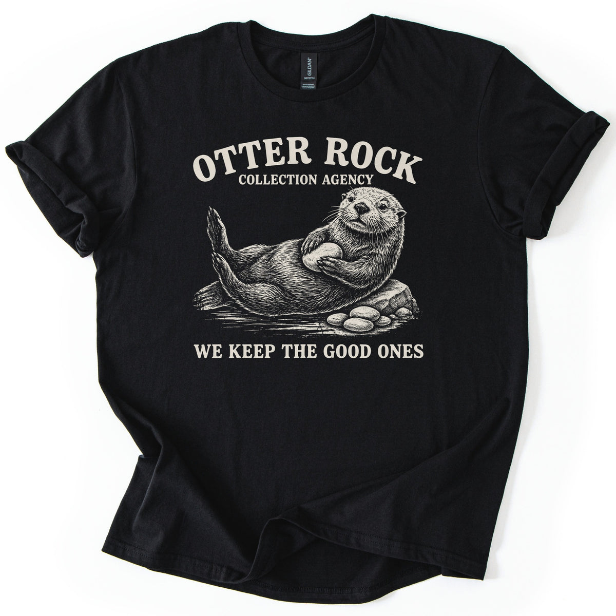 Otter Rock Collection Agency Comfort Colors T-shirt in Black, laid flat. A cream engraving-style sea otter leans back on a rock clutching a round stone, pebbles beside it, below the words Otter Rock Collection Agency and above We Keep the Good Ones. Sleeves cuffed, on a white surface.