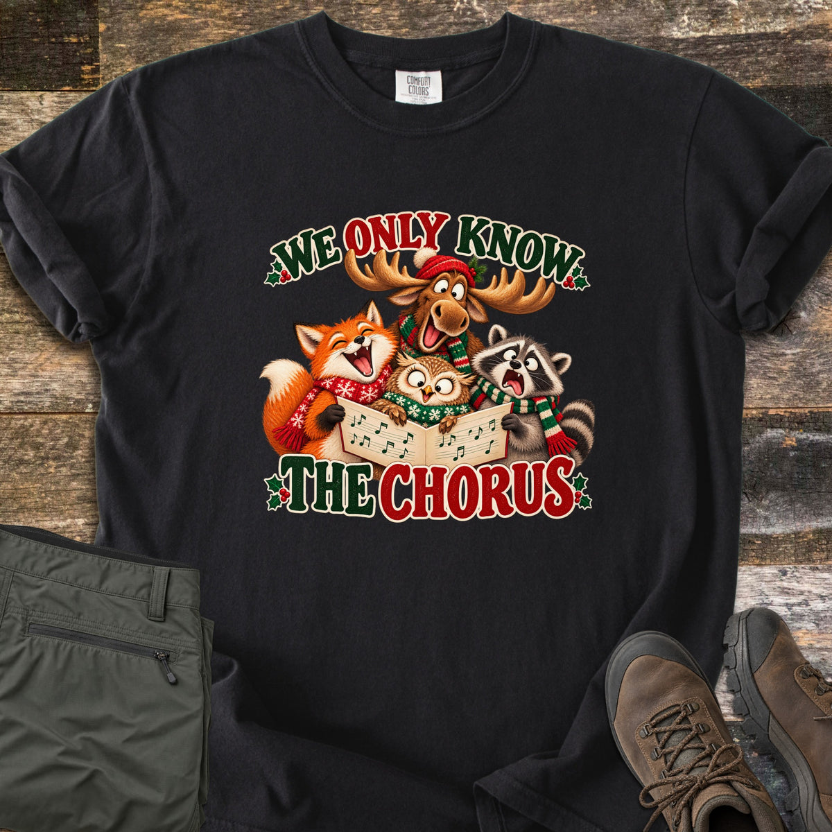 We Only Know the Chorus Comfort Colors T-shirt in Black, laid flat. A cartoon moose in a red hat, a fox, an owl and a raccoon in scarves sing from a songbook between the words We Only Know The Chorus. Shown on wood boards with green pants and brown boots.
