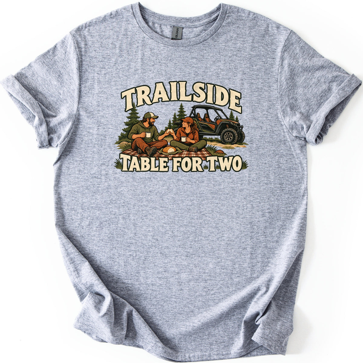 Trailside Table for Two Comfort Colors T-shirt in Sport Grey, laid flat. The words Trailside and Table for Two frame a couple in caps picnicking on a plaid blanket with mugs and a loaf of bread, a side-by-side UTV and pine trees behind them. Laid on a white surface.