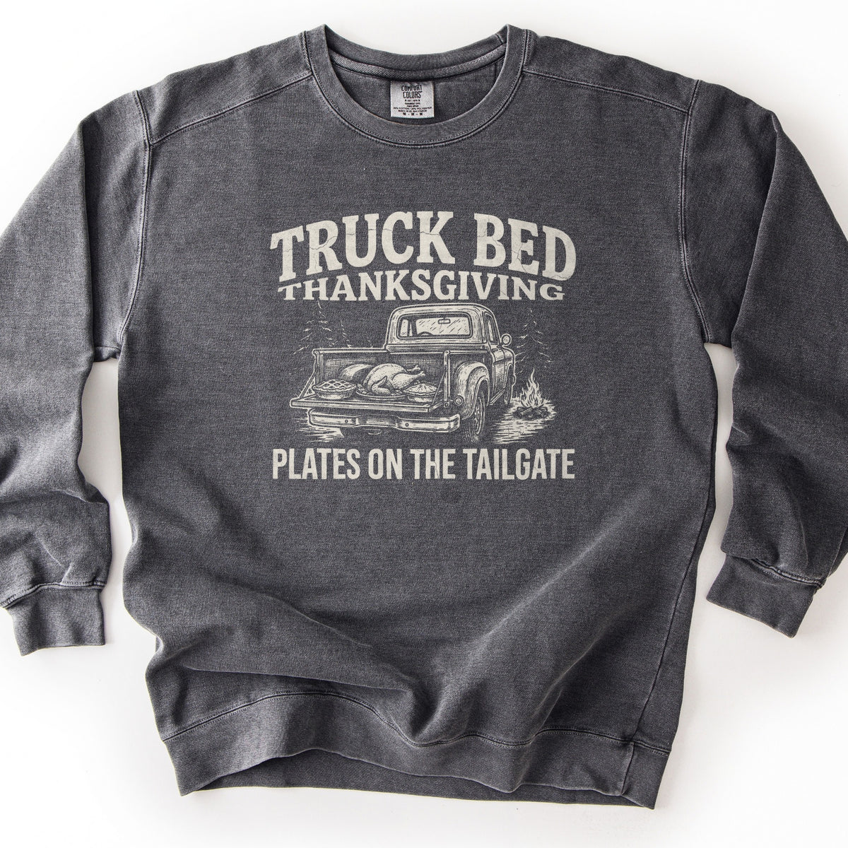 Truck Bed Thanksgiving Comfort Colors crewneck sweatshirt in Pepper, laid flat. Cream distressed print of an old pickup truck with a roast turkey and pies on its open tailgate, pine trees and a campfire nearby, between the words Truck Bed Thanksgiving and Plates on the Tailgate. Shown on a white background.