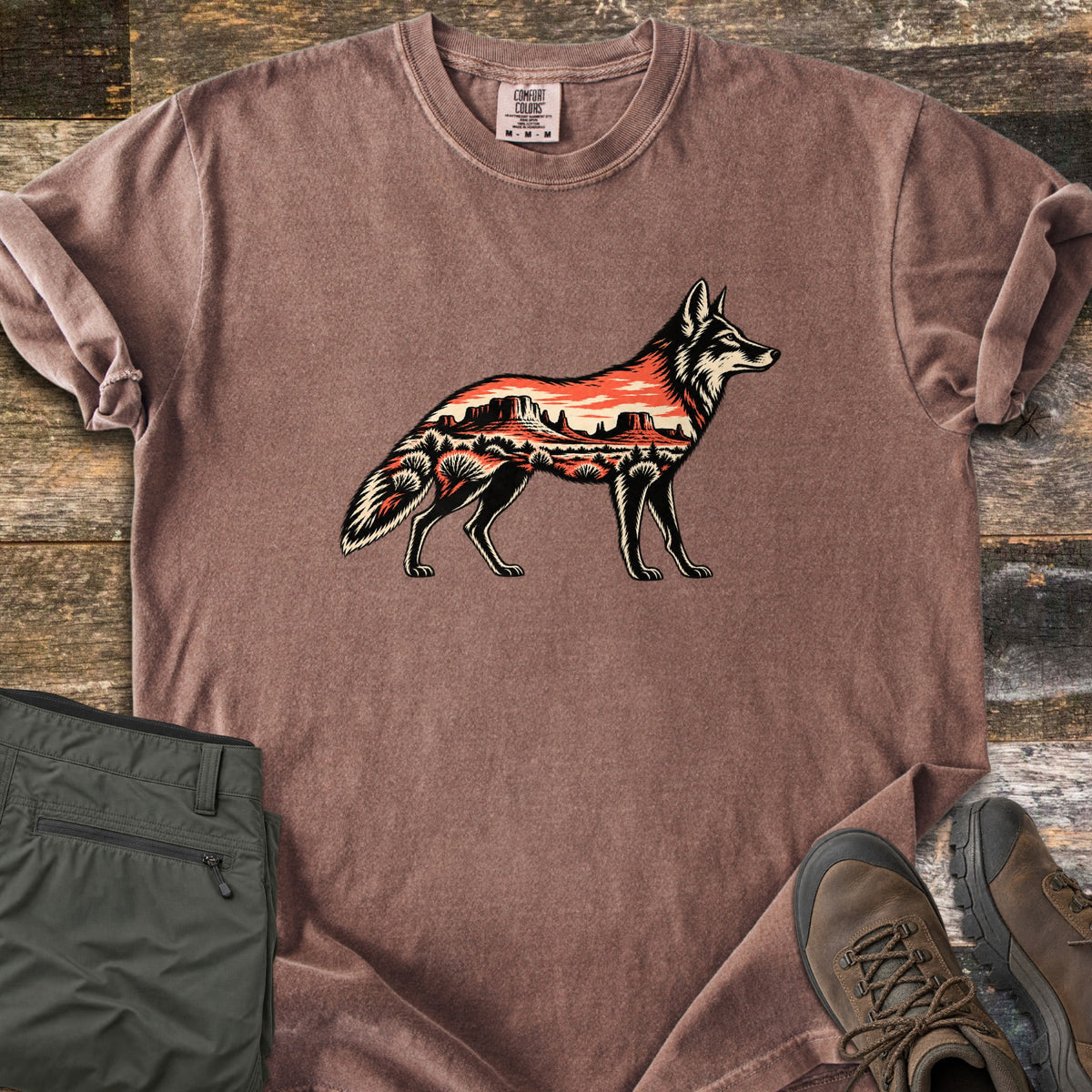 Desert Mesa Coyote Comfort Colors T-shirt in Espresso, laid flat. A side-view coyote silhouette filled with a red desert mesa landscape, sunset sky and shrubs, with no words. Sleeves rolled on rustic wood, beside folded olive hiking pants and brown hiking boots.